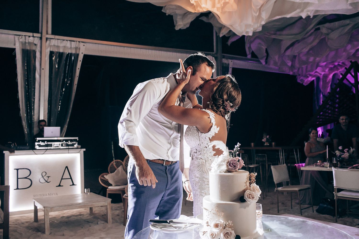 A couple shares their first dance at a wedding reception under purple mood lighting and white decor.