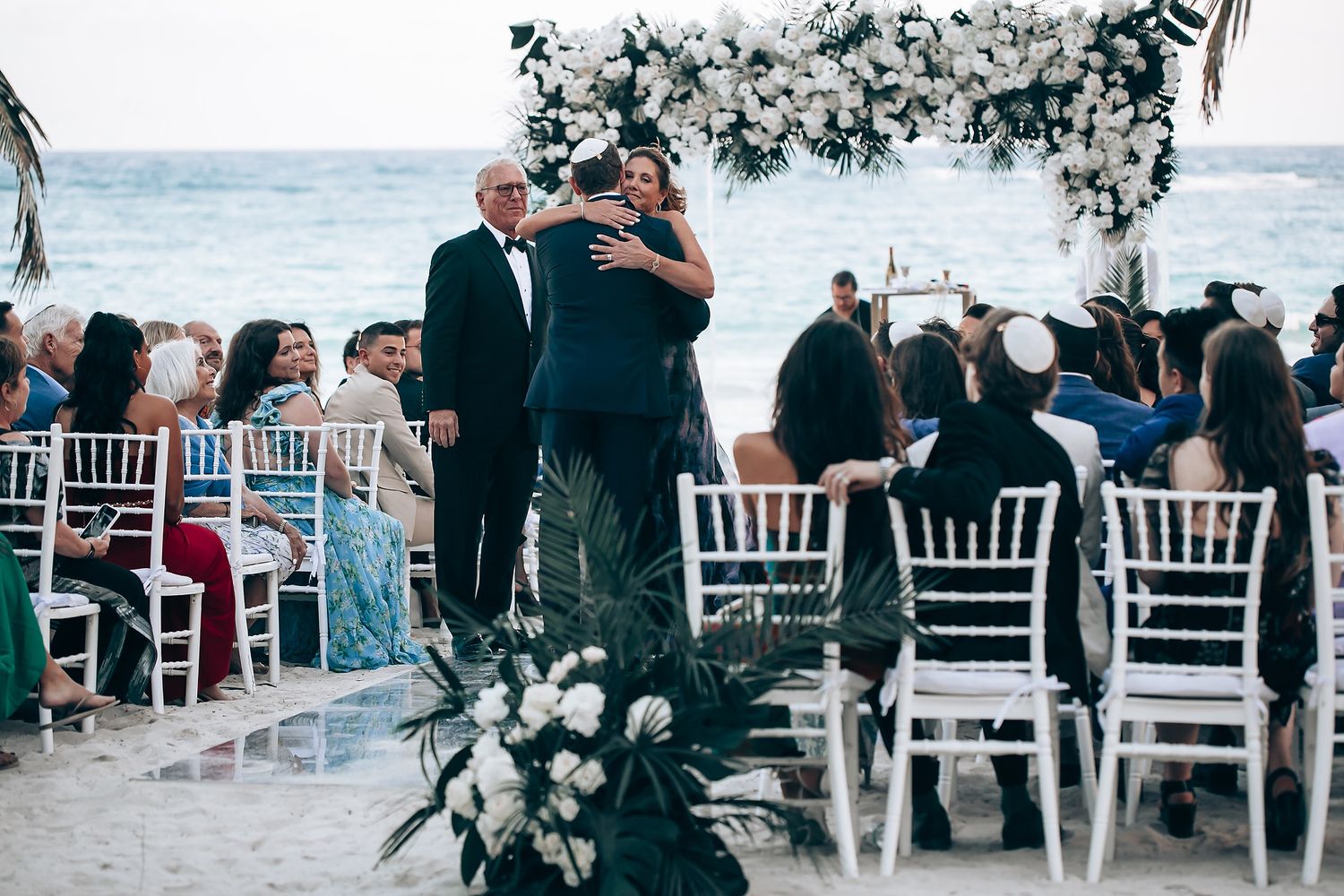 Guests seated in white chairs for an outdoor beach wedding ceremony with palm trees and ocean views at sunset.