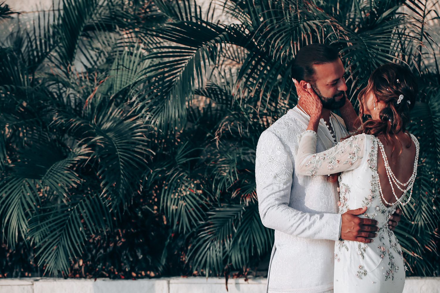 A romantic couple in white attire embraces tenderly among tropical palm trees during an intimate outdoor photo session.