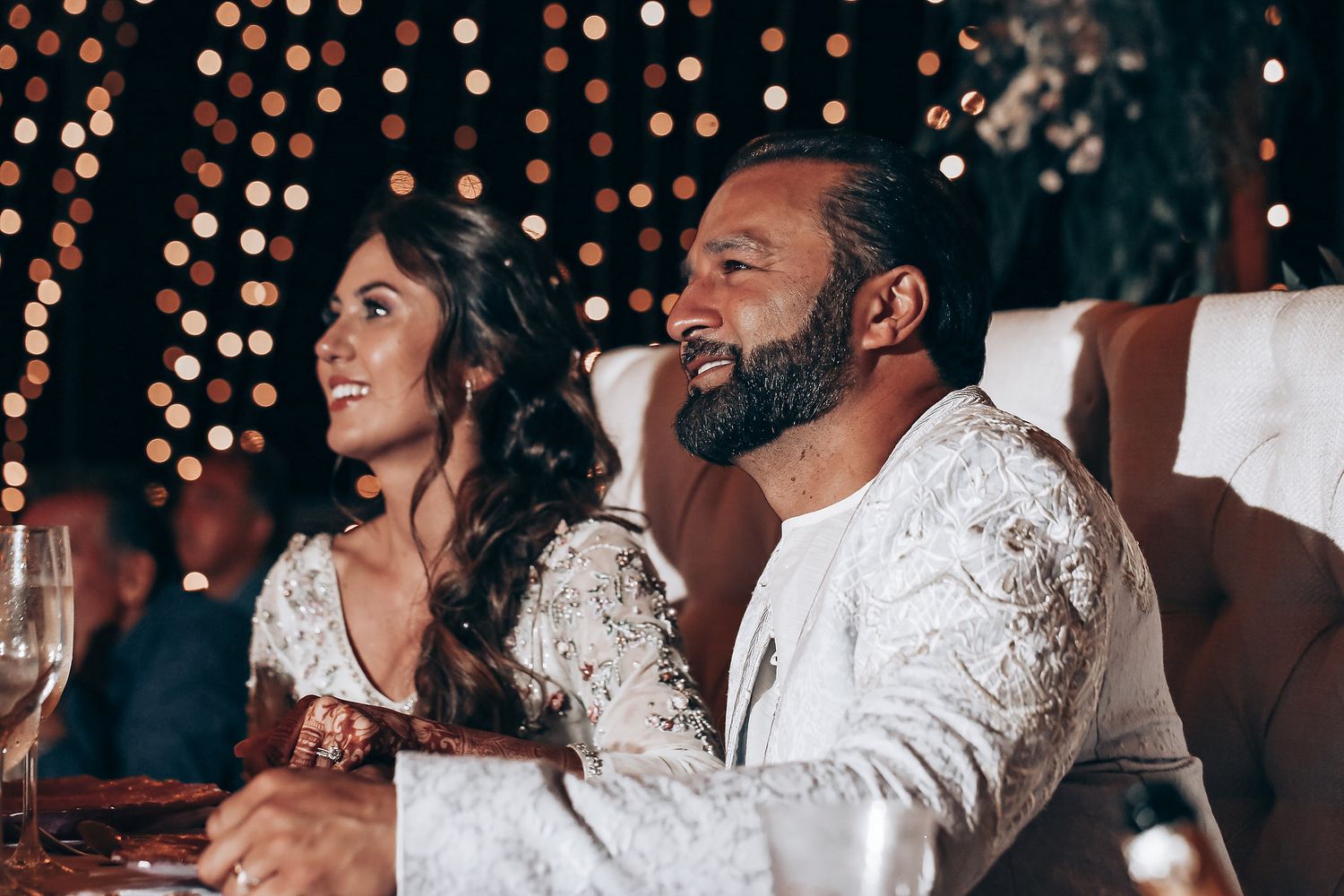 Wedding guests in white lace attire seated at tables during an evening reception with bokeh lights in background.