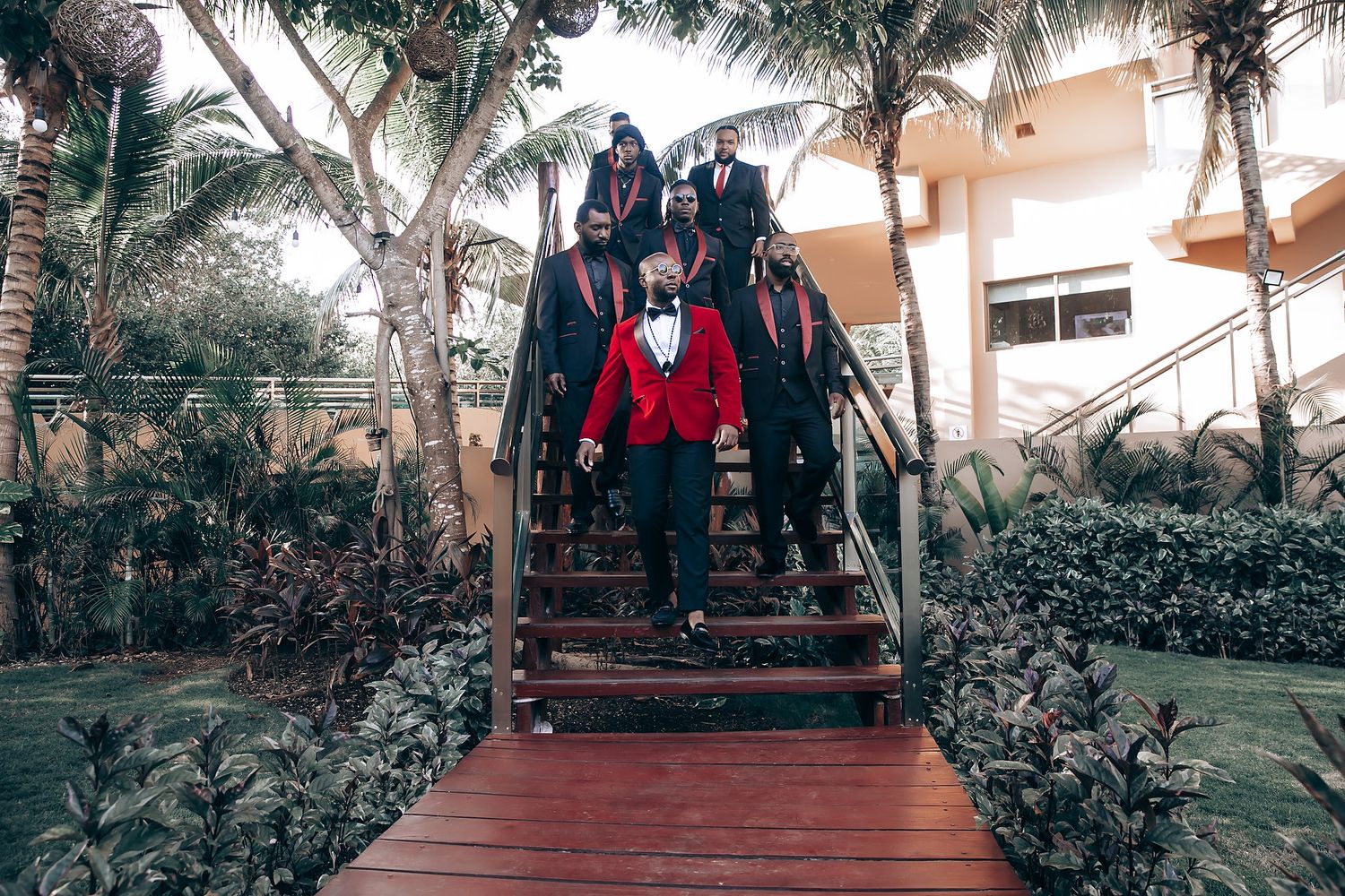Wedding party descending wooden stairs in a tropical garden setting with palm trees and lush landscaping.