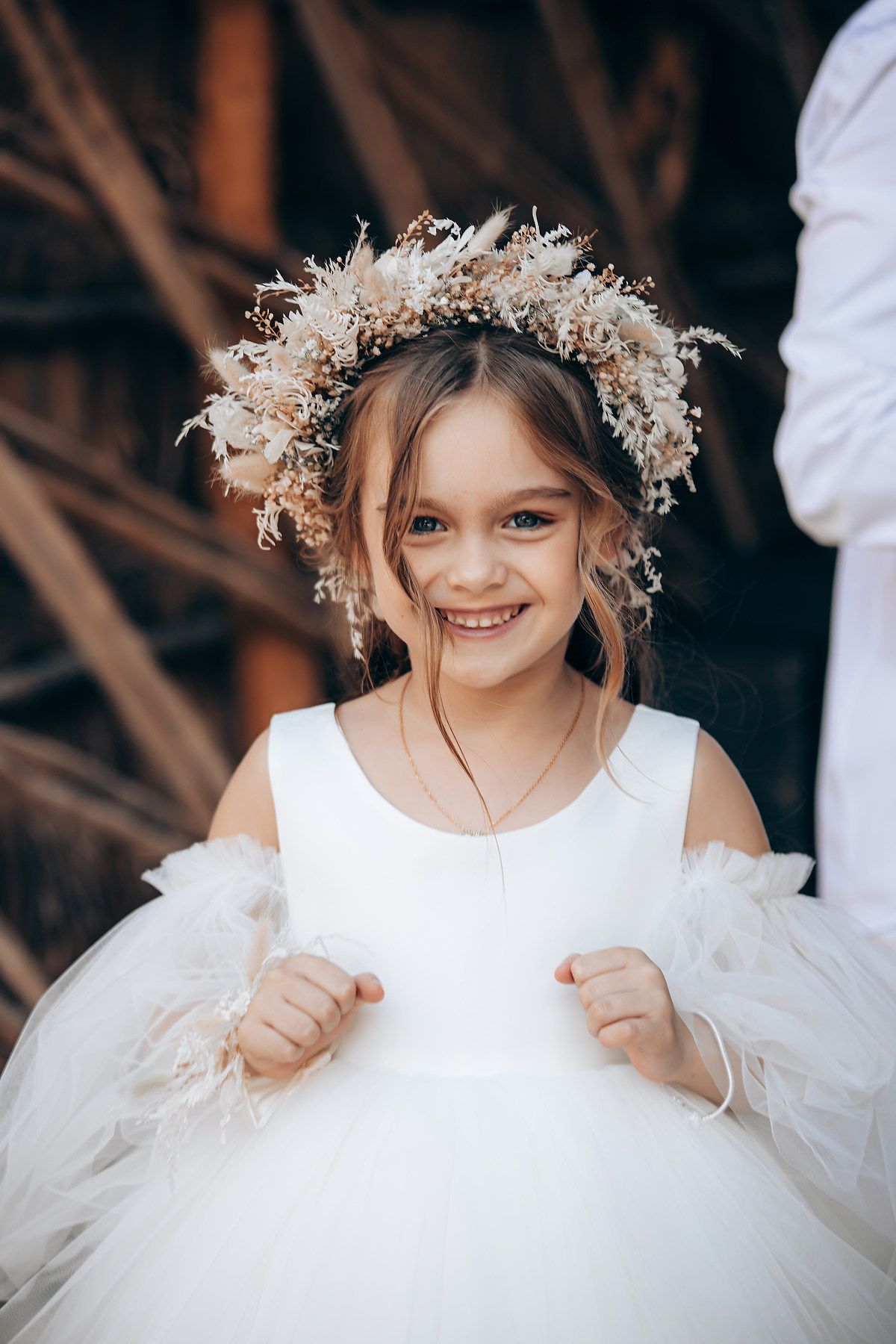 A flower girl in a white tulle dress wears a delicate dried floral crown at a wedding.