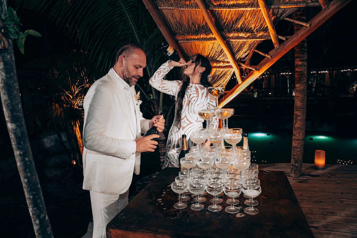 Champagne tower being carefully poured at an elegant evening wedding reception with warm amber lighting.