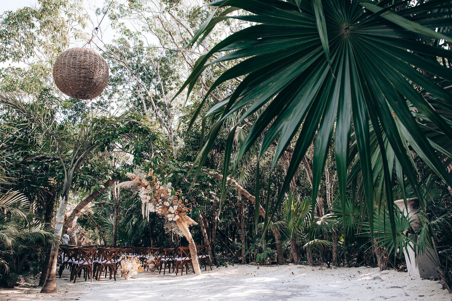 Rustic outdoor wedding venue in a tropical garden setting with palm trees and hanging rattan lantern decor.