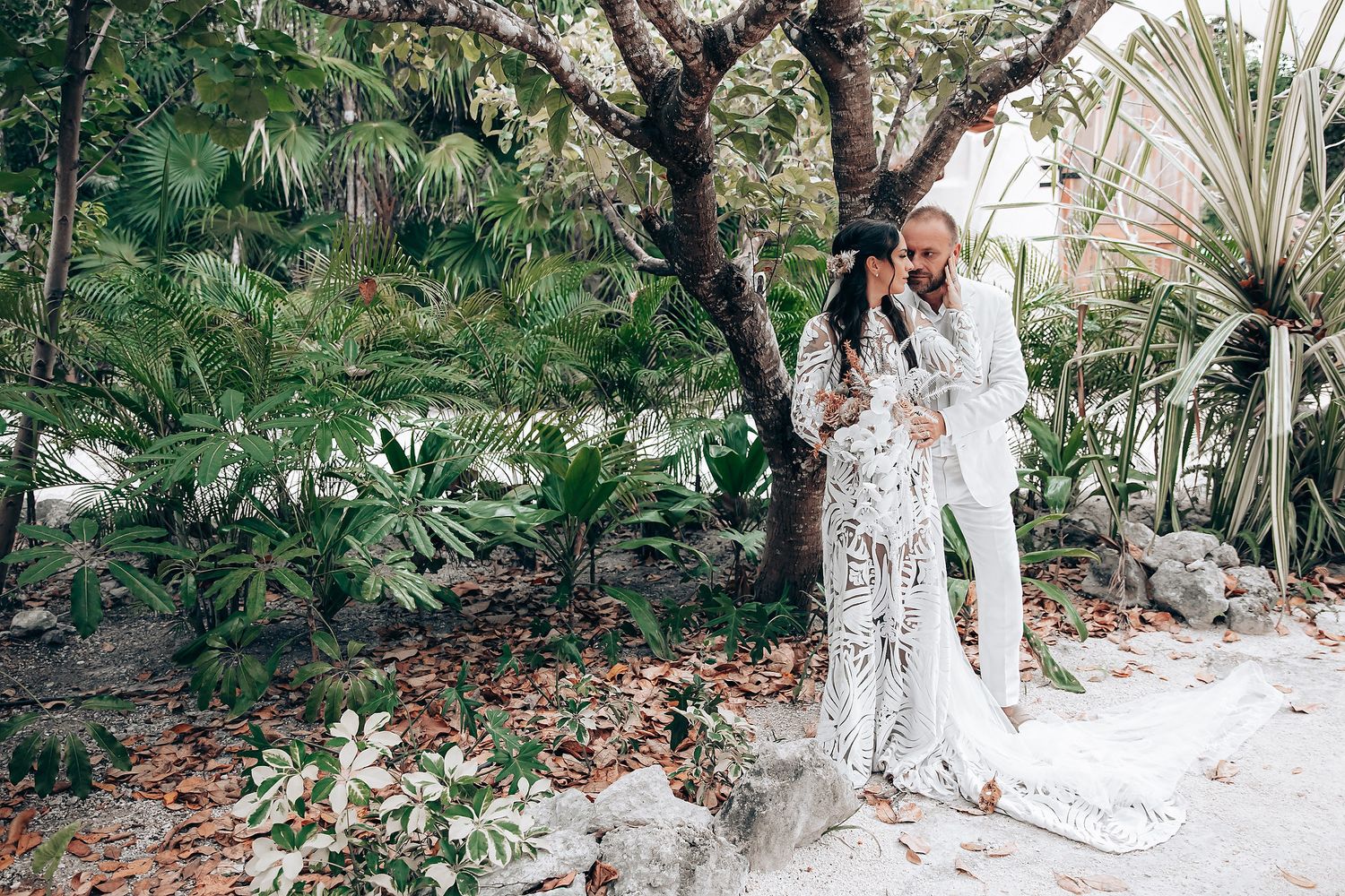 A couple embraces under a tropical tree in a lush garden setting wearing elegant white attire.