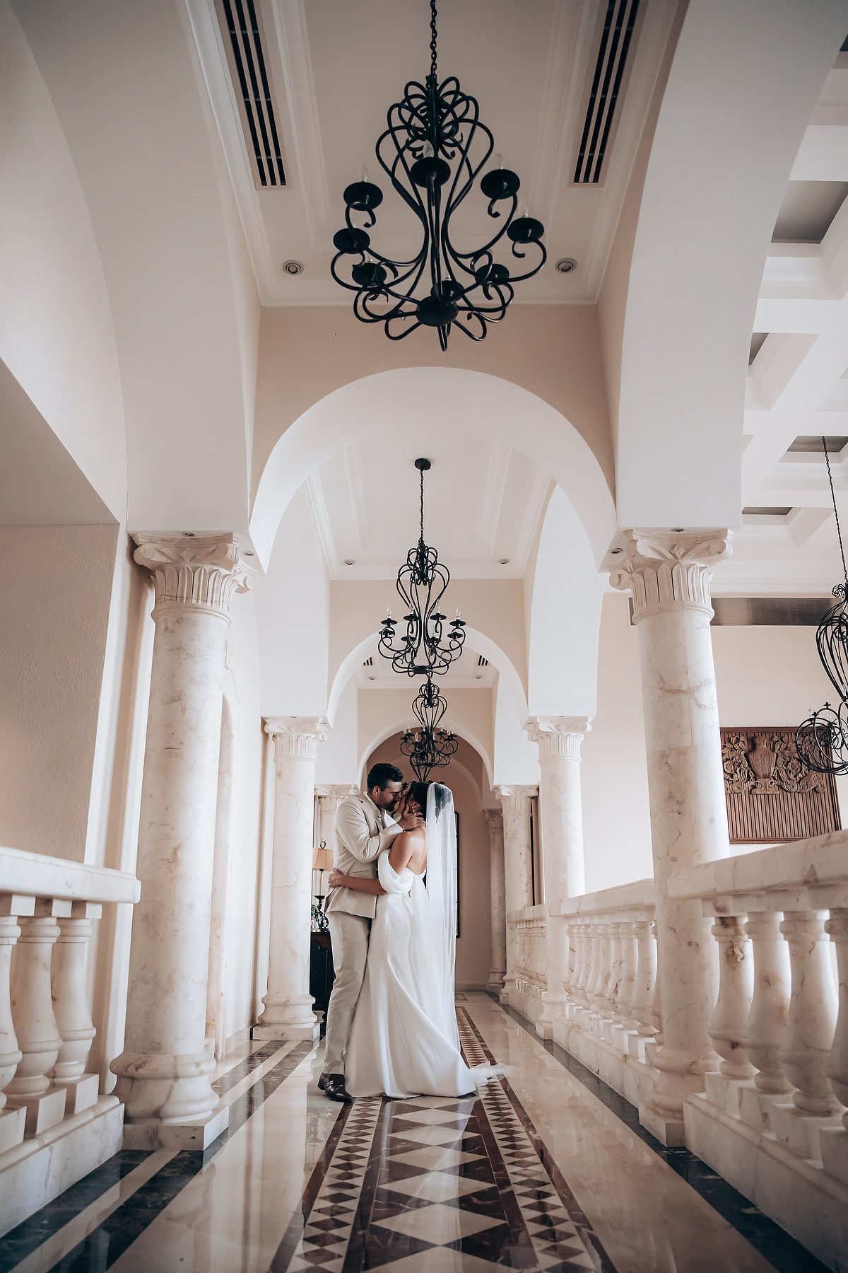Elegant white hallway with arched columns and wrought iron chandeliers creates a romantic setting for a wedding photo shoot.