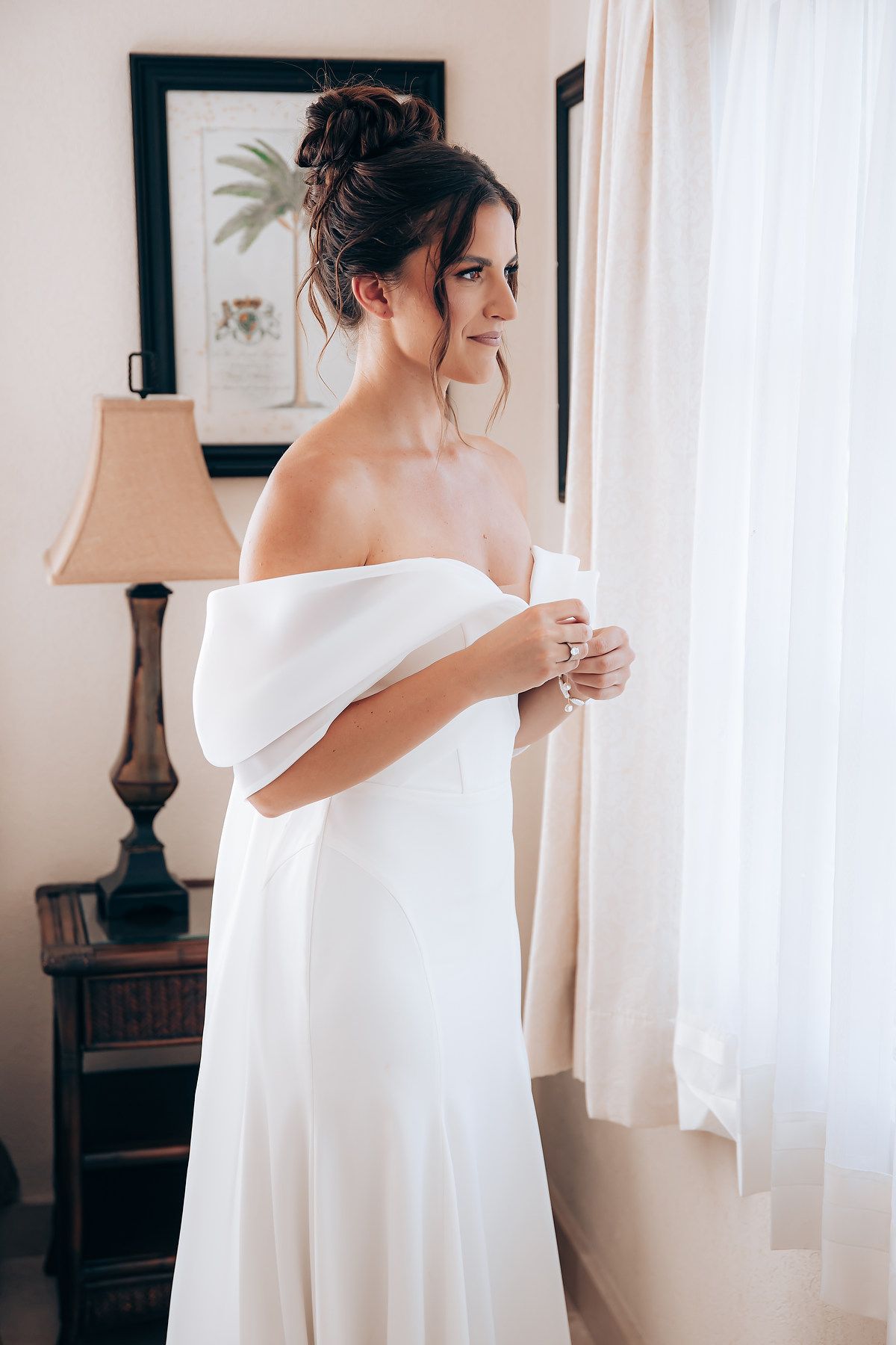A woman in an elegant white off-shoulder dress stands by a window in a softly lit room with delicate decor.