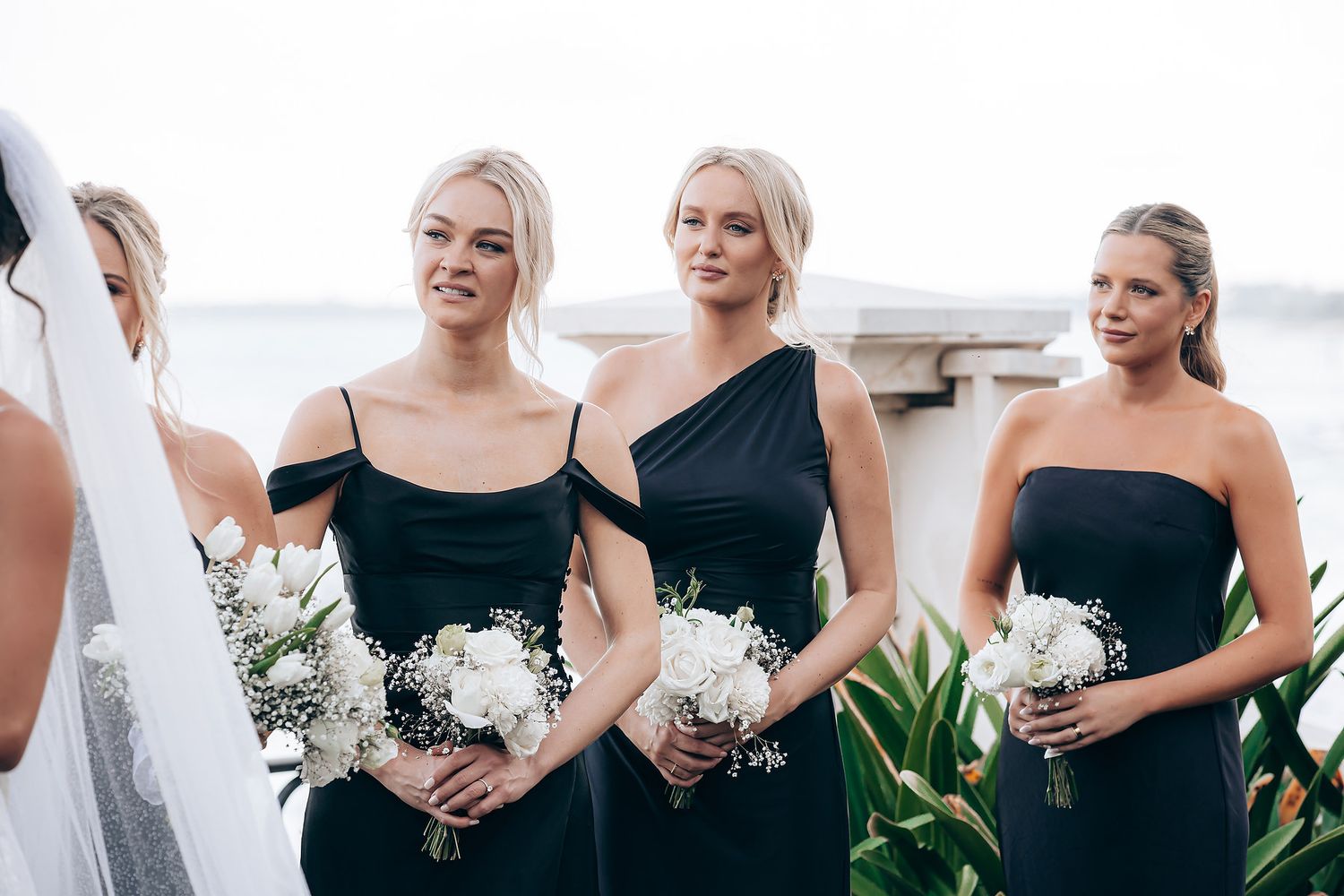 Bridesmaids in elegant black dresses hold white floral bouquets while standing together at a wedding venue.