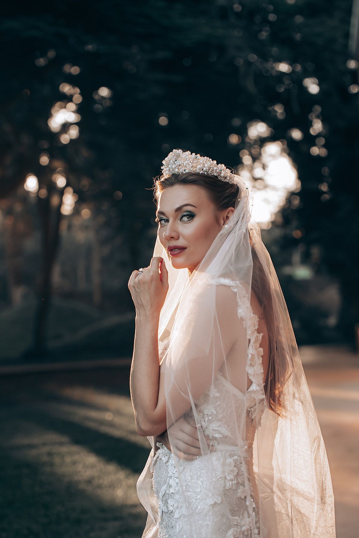 A bride in a white lace wedding dress and veil poses against a sunlit outdoor background during golden hour.