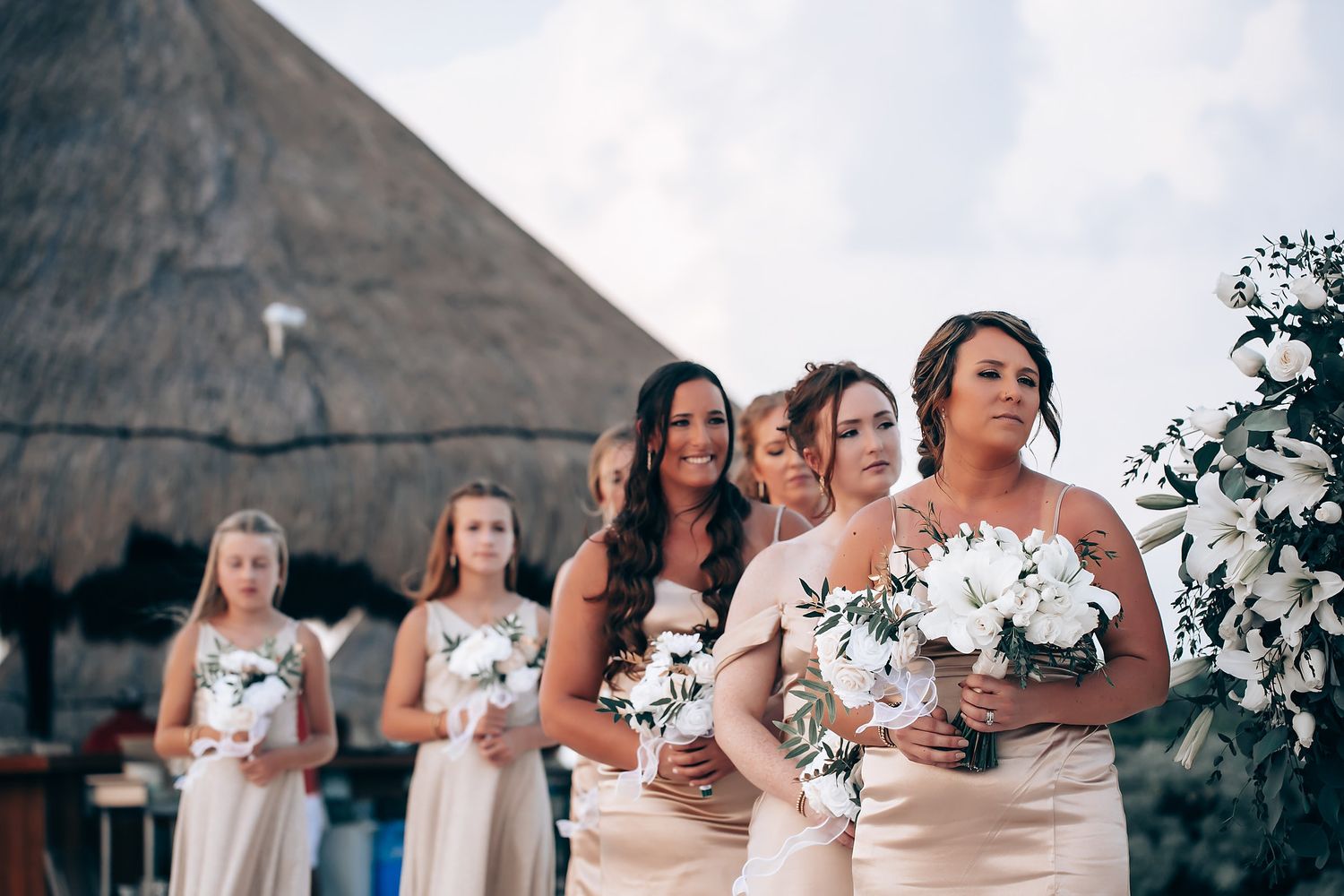 Wedding party holding white floral bouquets against tropical beach backdrop.