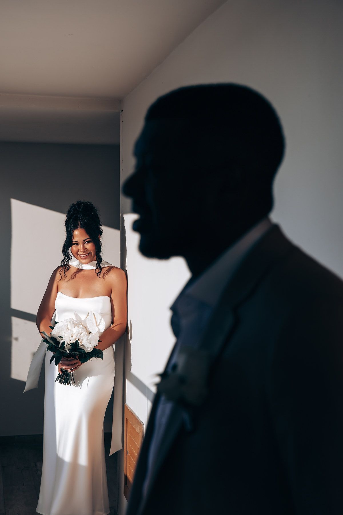 Silhouetted figures in intimate wedding moment with dramatic window lighting.