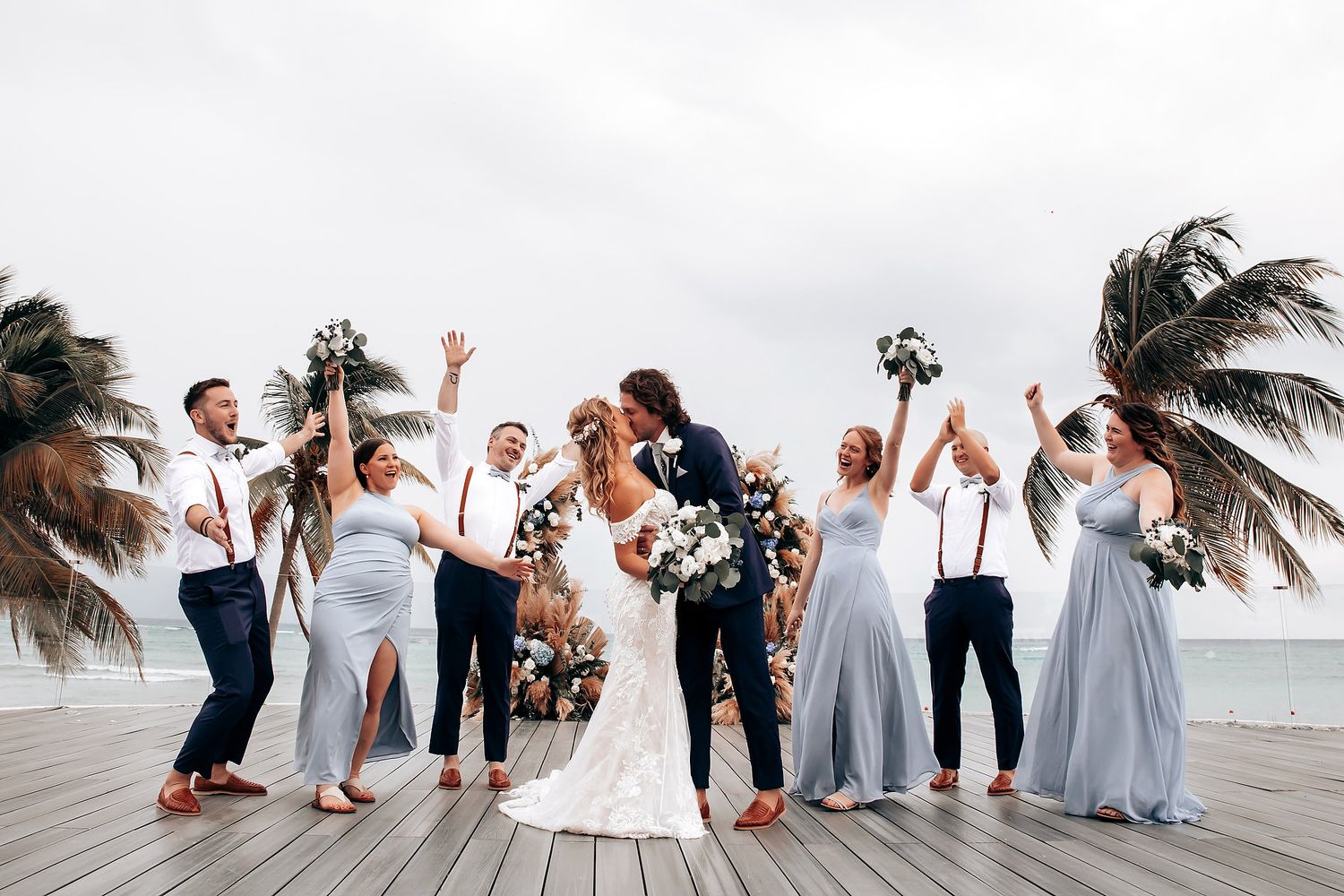 Wedding party jumping and celebrating on a beachfront deck with palm trees in the background.