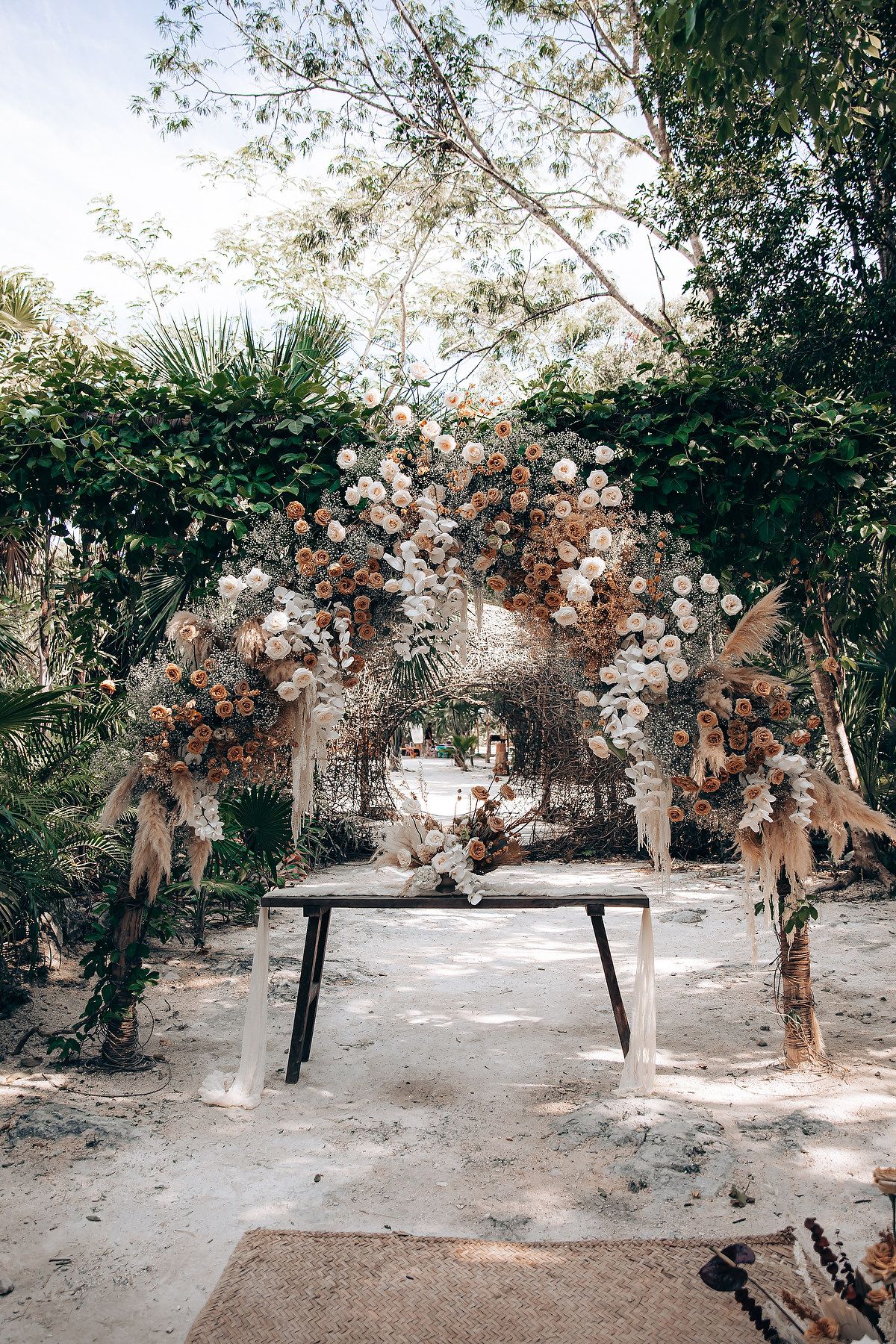 Wedding ceremony setup with floral arch and white chairs in a tropical garden setting.