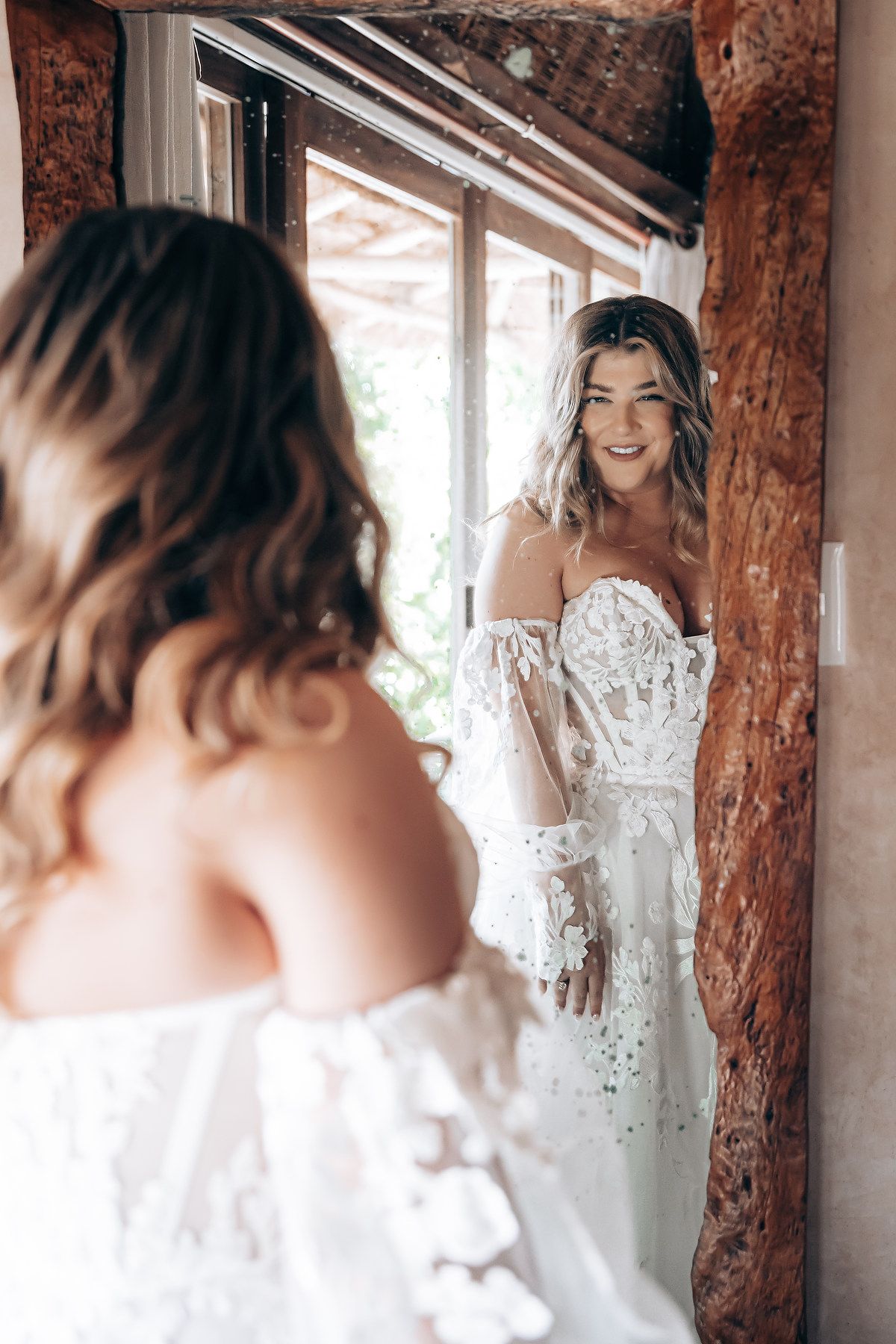 Bridal preparation moment captured in mirror reflection with delicate off-shoulder wedding gown.