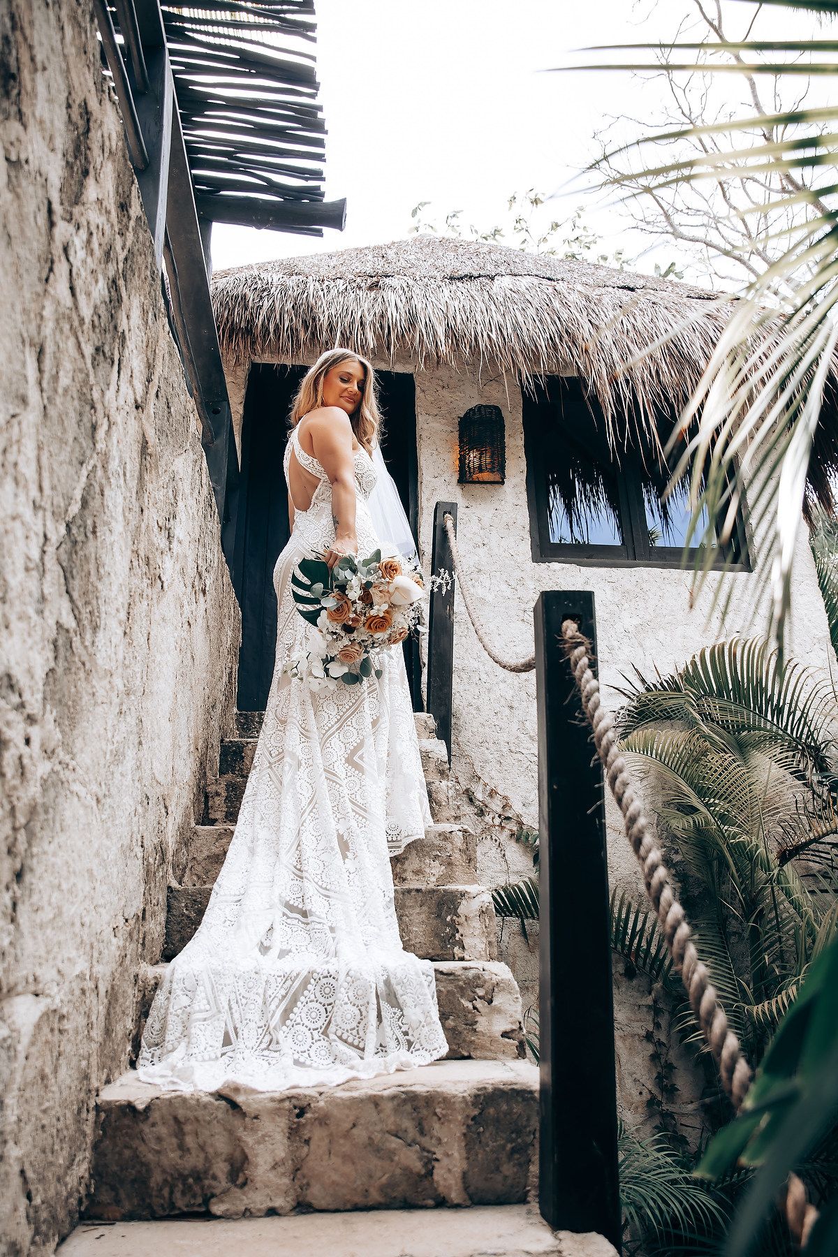 A wedding dress flows elegantly against a rustic backdrop with palm trees and thatched roof huts in a tropical setting.