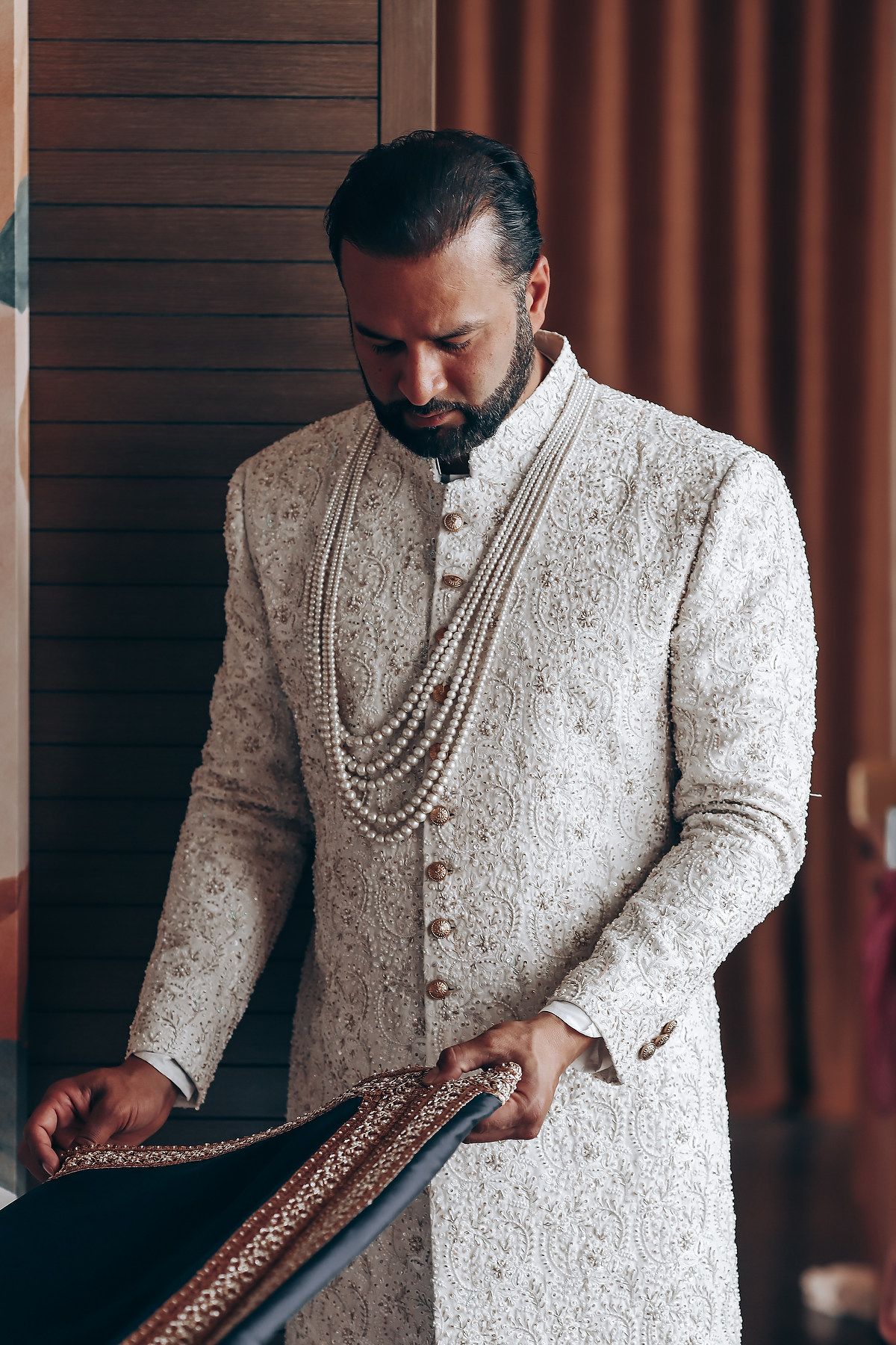 A person adjusts traditional Indian wedding attire in an ornate white sherwani with pearl details.