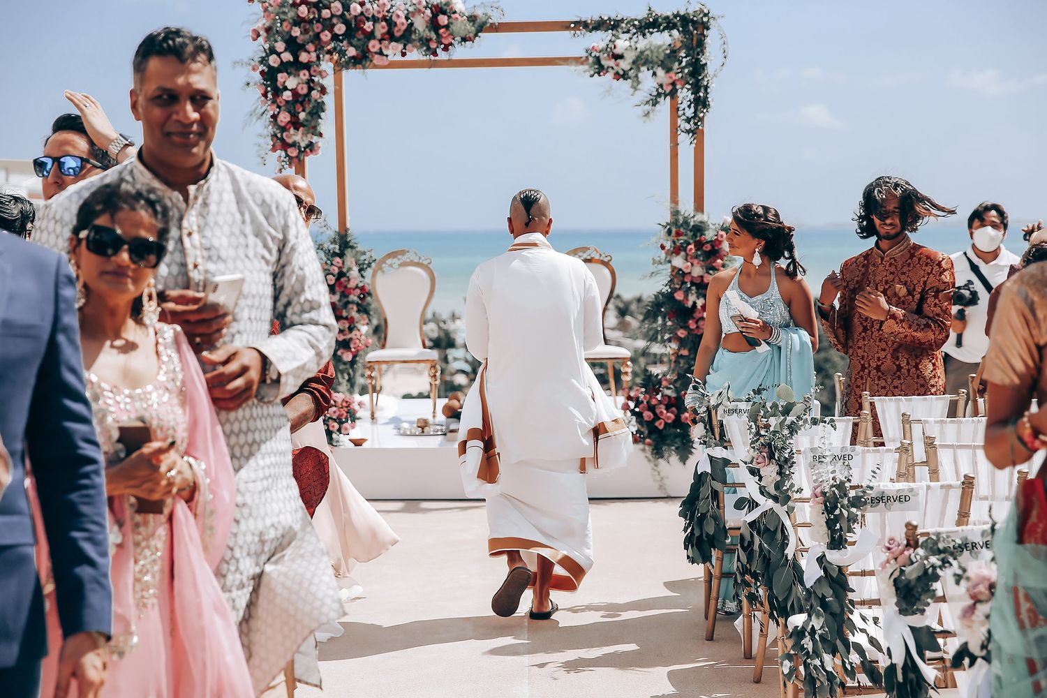 Wedding ceremony setup with floral arch and white chairs on a sunny beach.