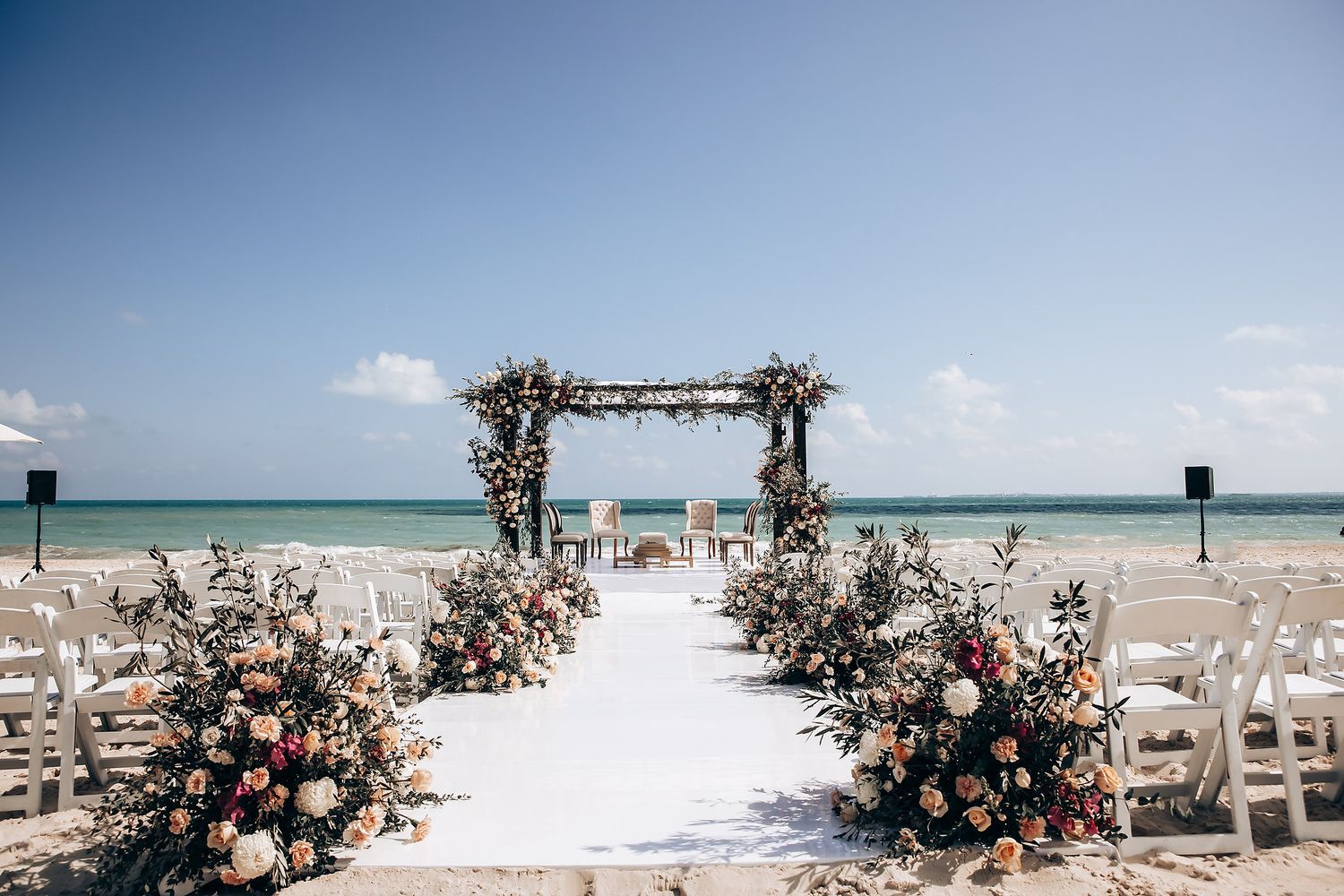 A beach wedding ceremony setup with white chairs and floral decorations under a wooden arch by the turquoise ocean.