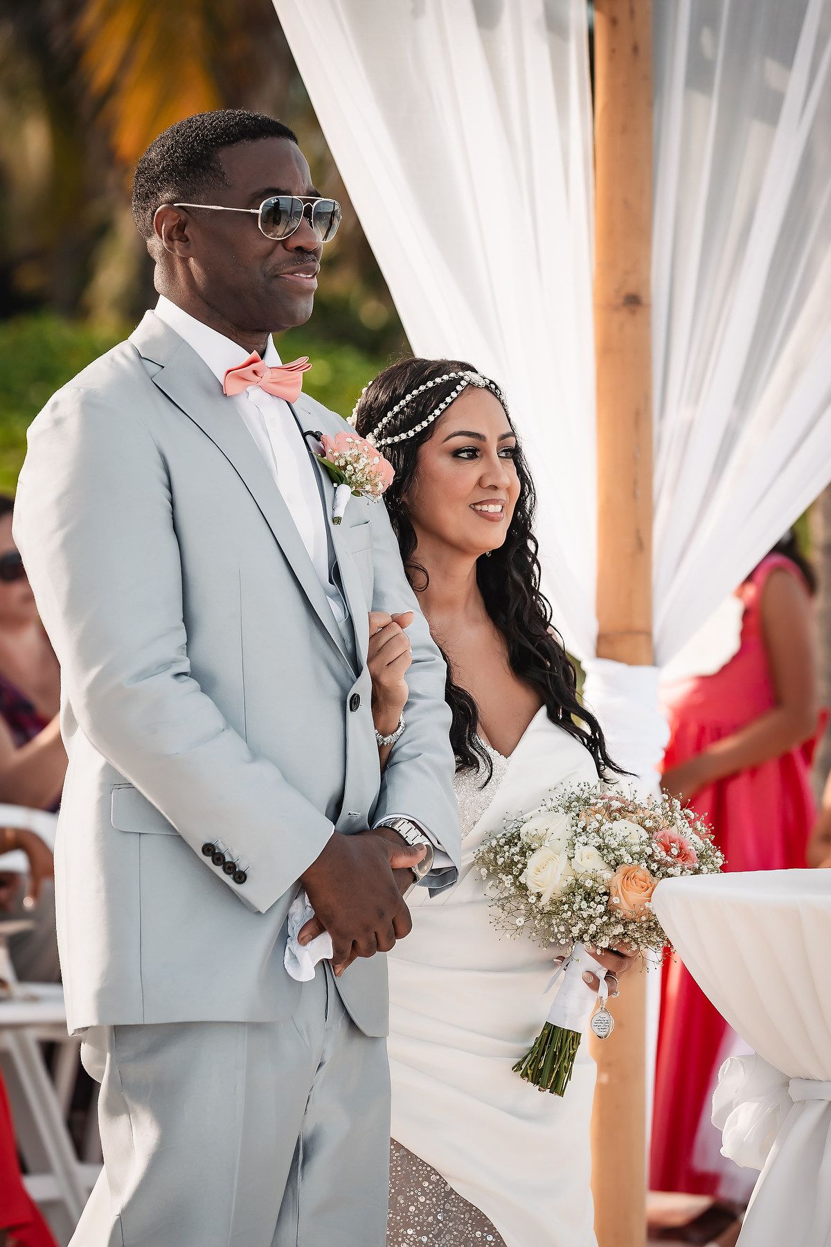 A couple stands together during their beach wedding ceremony with palm trees and white decor in the background.