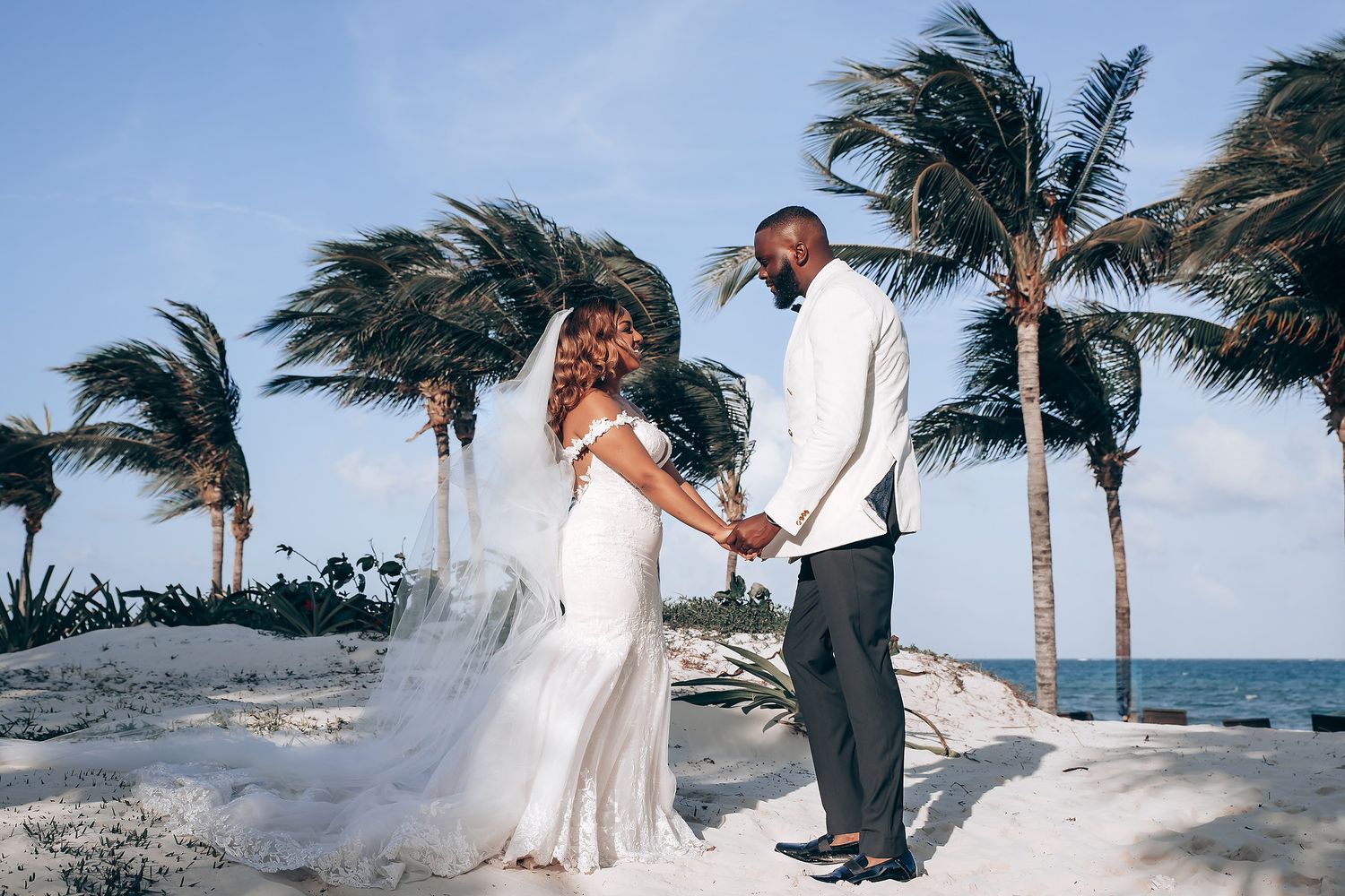 Couple in wedding attire poses on a windy beach with palm trees swaying in the tropical breeze.