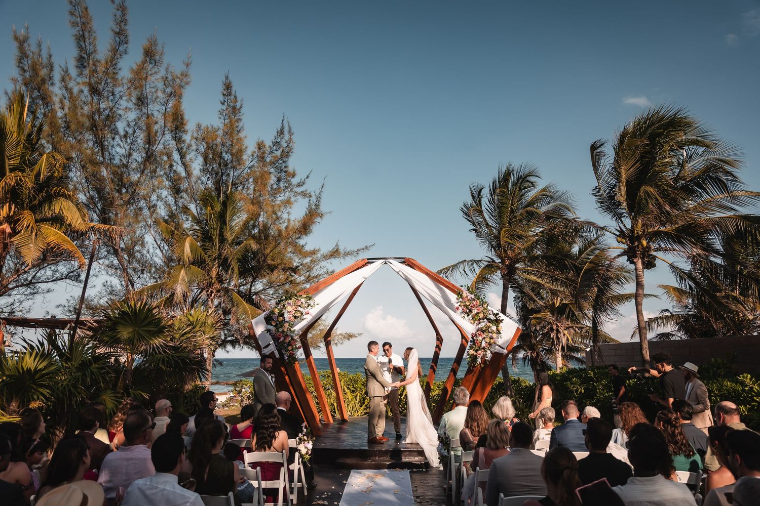Tropical beach wedding setup with wooden architectural structure and white flowing fabric.