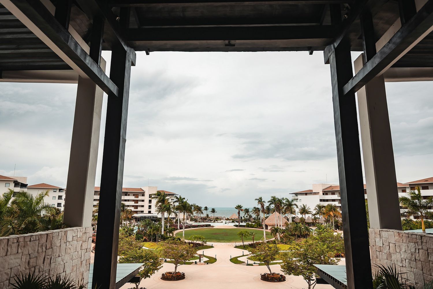 Luxury resort courtyard with palm trees, manicured gardens, and ocean view framed by elegant wooden columns under cloudy sky.