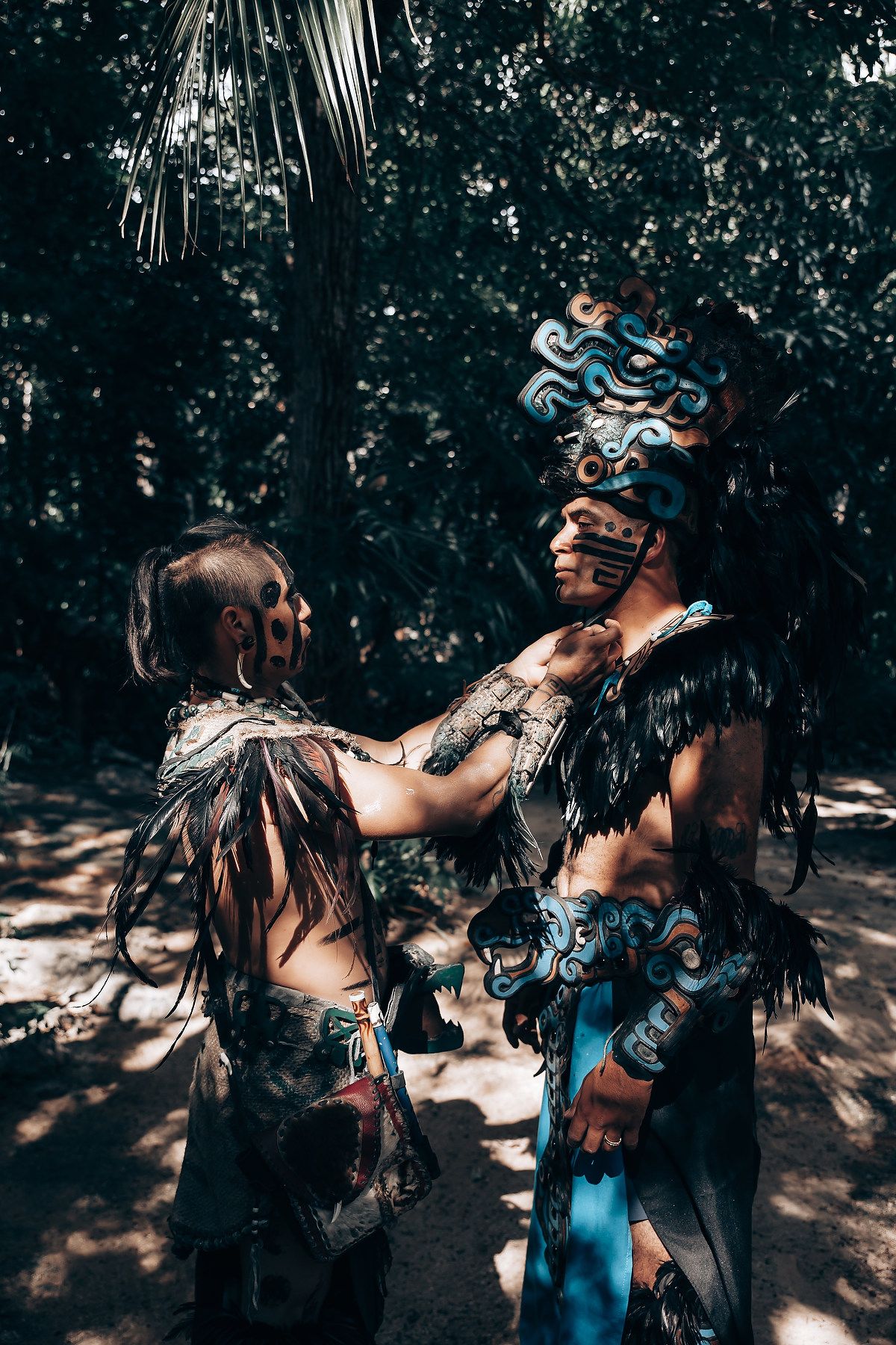 Two performers in elaborate Mayan costumes with face paint and feathered headdresses interact in a lush forest setting.