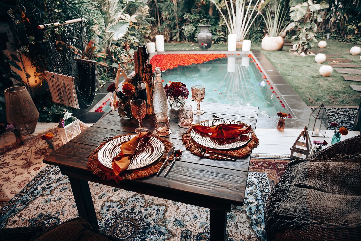Romantic outdoor dining table set with elegant red and gold tableware beside a pool surrounded by lush tropical gardens.