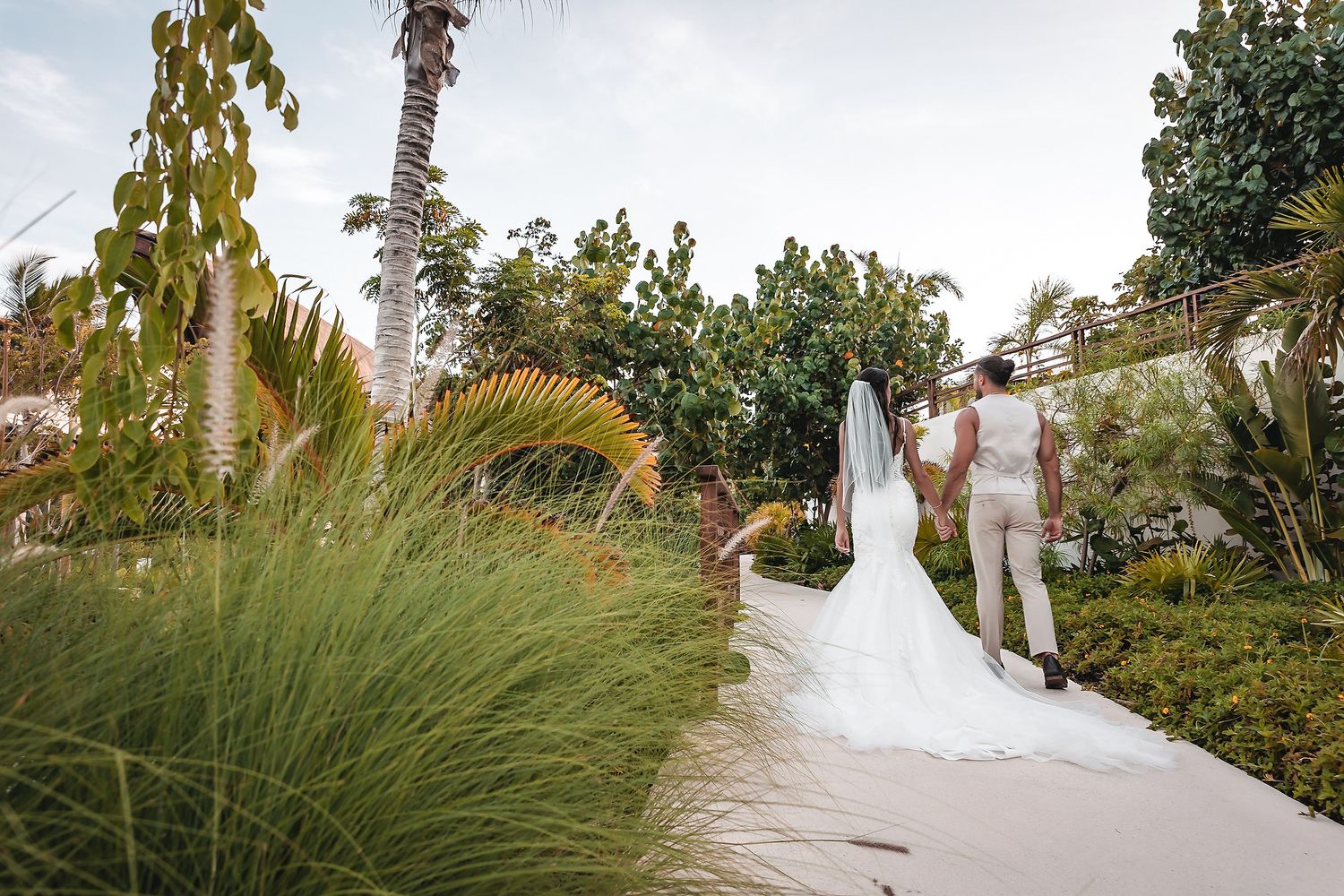 Bride and groom walking hand in hand through lush tropical garden path on their wedding day.
