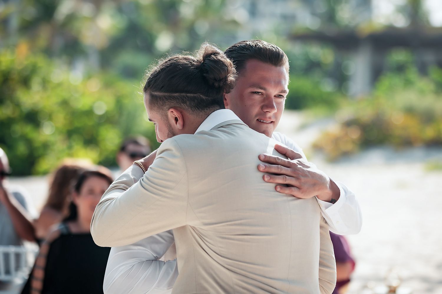 Two men in formal attire share an emotional hug at an outdoor wedding ceremony.