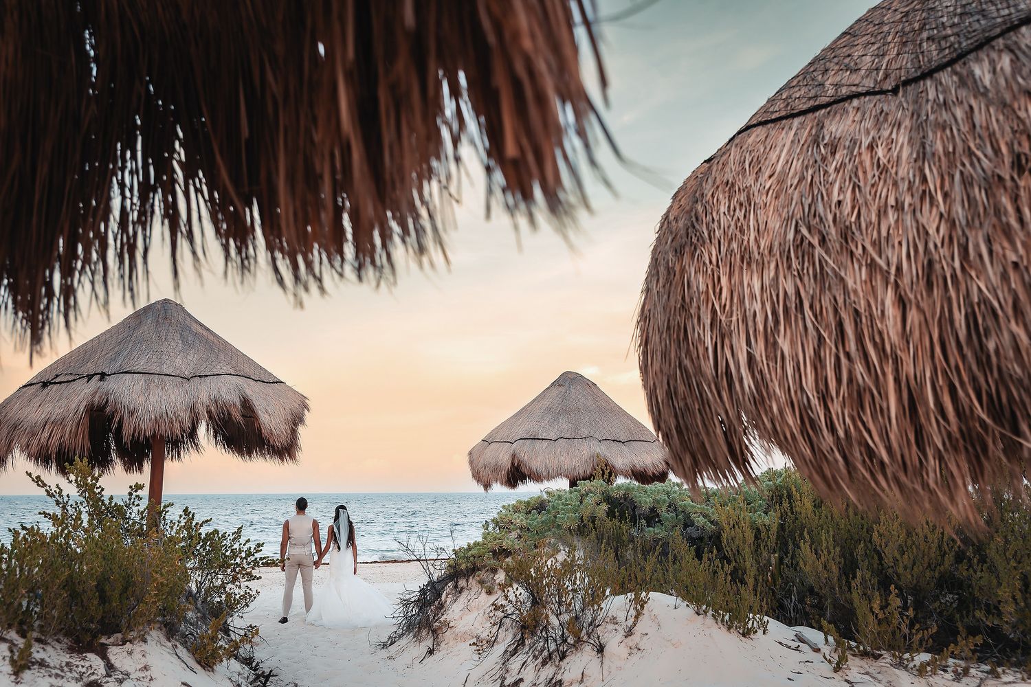 Couple walking on a white sand beach at sunset surrounded by thatched palapa umbrellas and tropical vegetation.