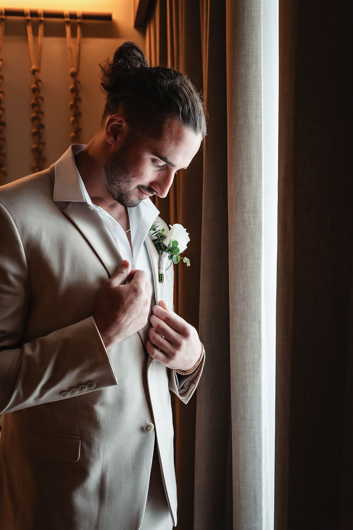 A smiling groom in a beige suit adjusts his white boutonniere while standing near a curtained window.