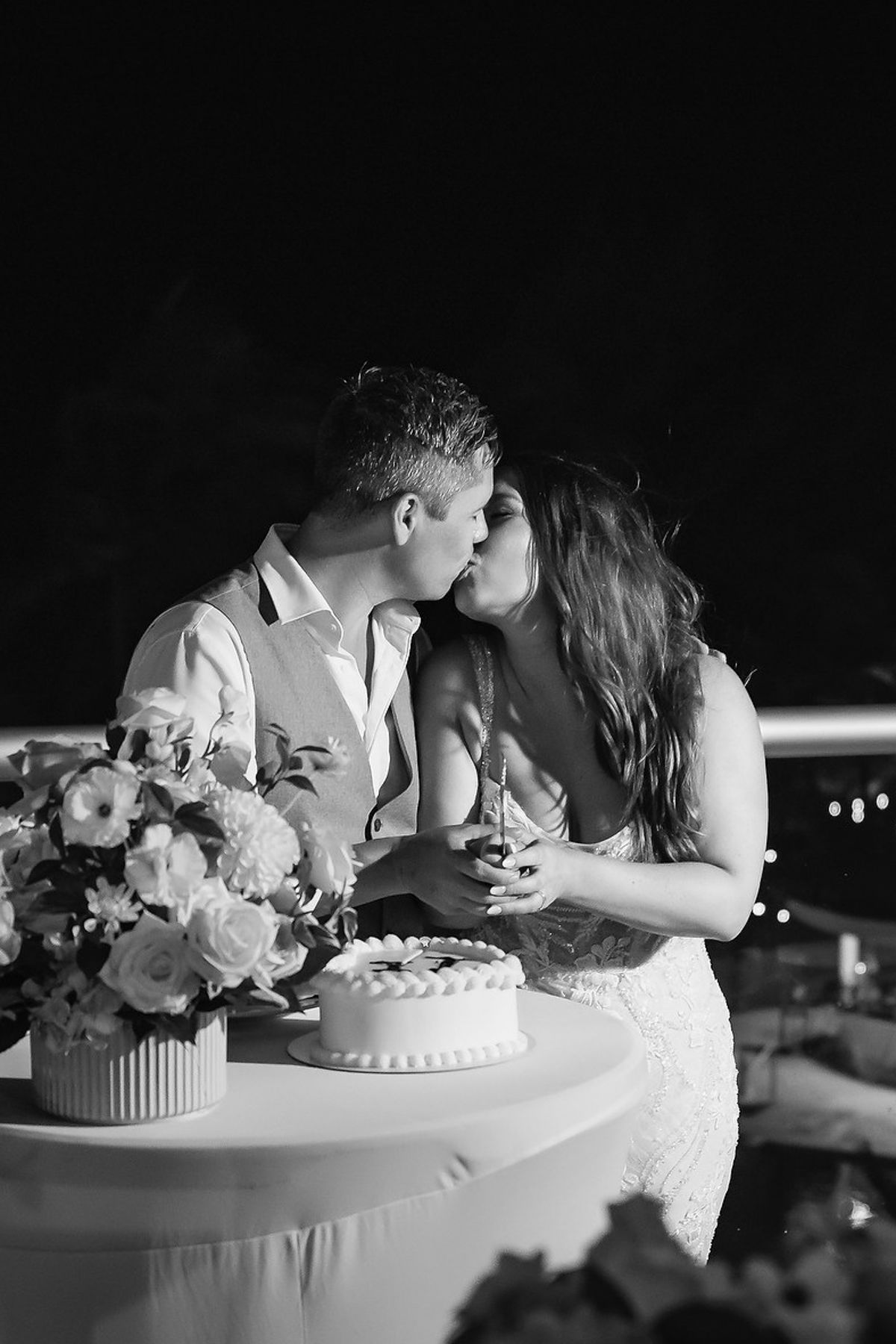 Black and white photo of a bride and groom sharing a kiss beside their wedding cake at an outdoor evening reception.