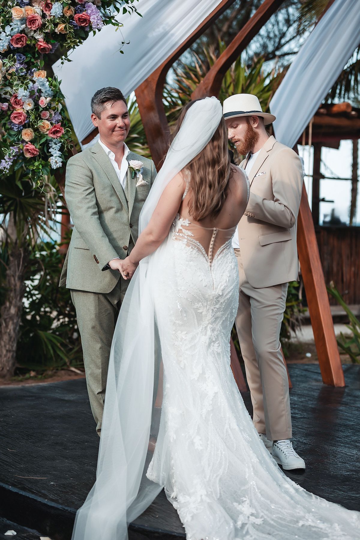 Bride in lace gown with veil exchanges vows with groom in tan suit at outdoor wedding ceremony with floral arch.