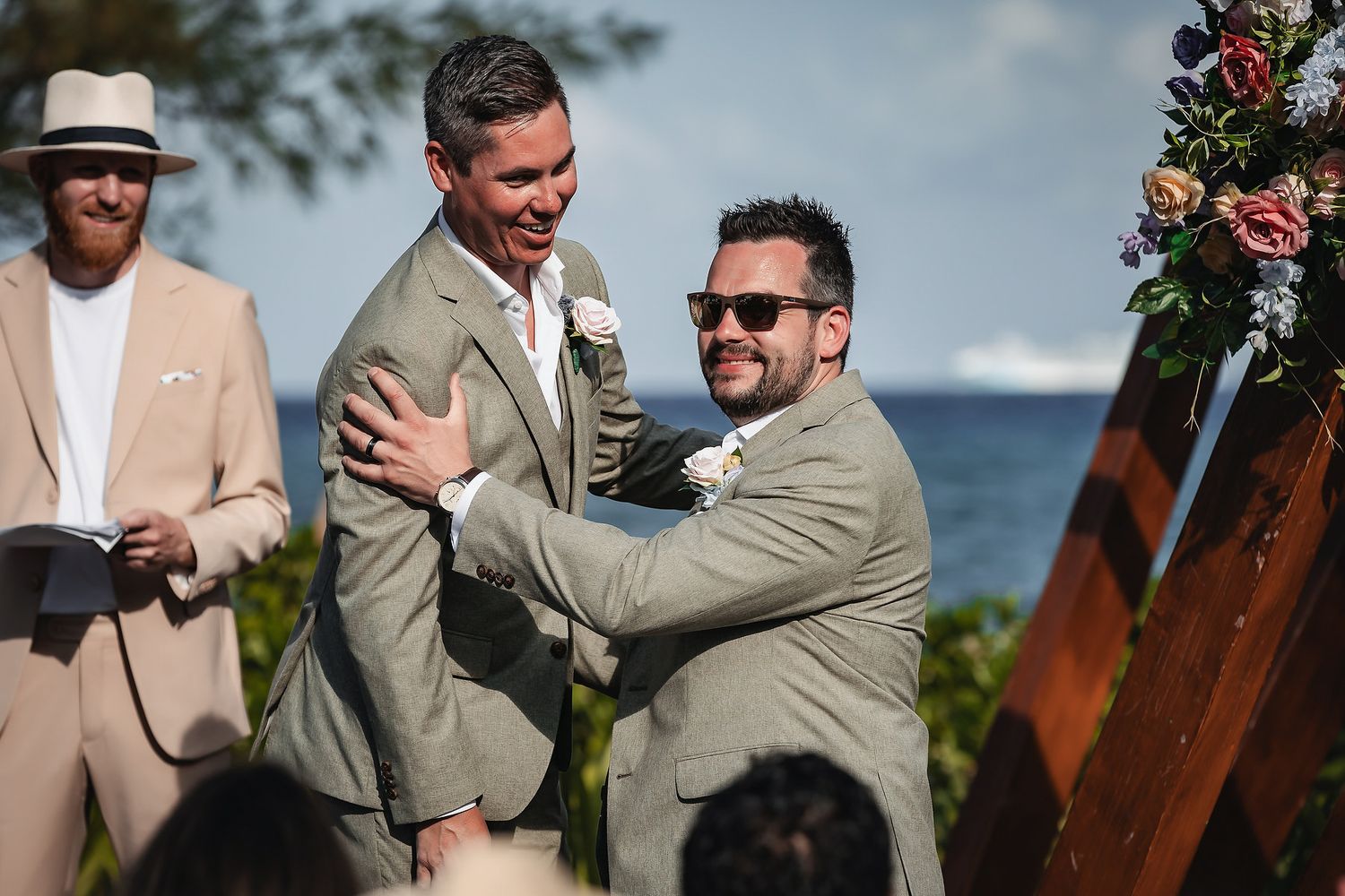 Two grooms share a joyful embrace at an outdoor beach wedding ceremony with ocean views.