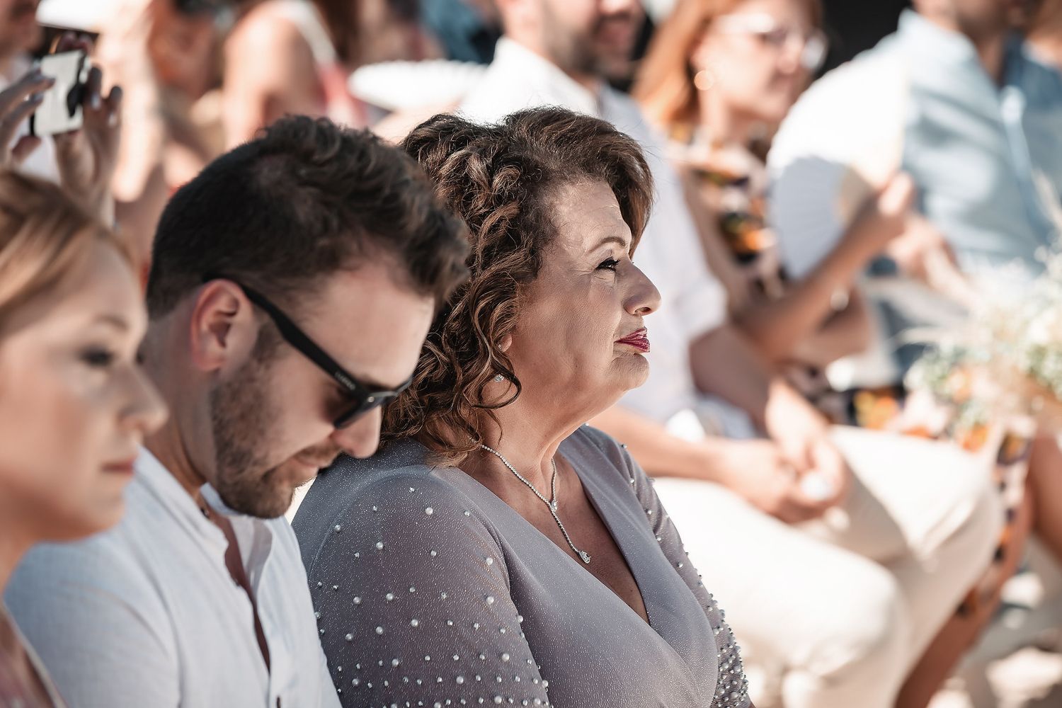 Wedding guests watch emotional outdoor ceremony, woman in grey dress seated beside man in sunglasses.