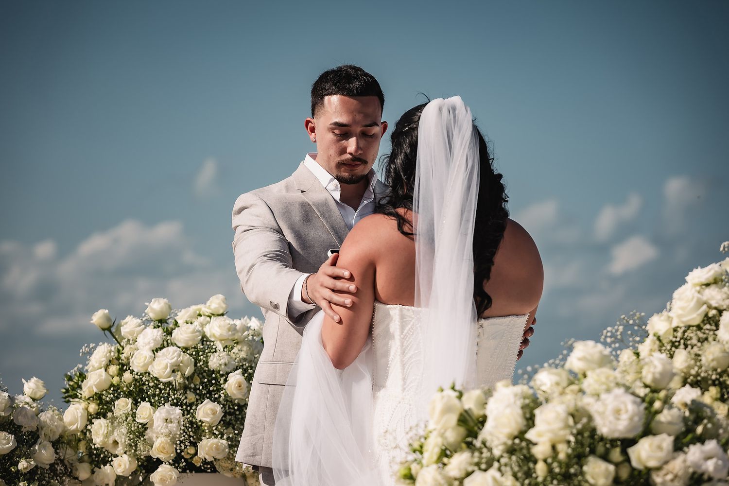 Bride and groom share a romantic embrace at an outdoor wedding ceremony surrounded by white floral arrangements under a blue sky.