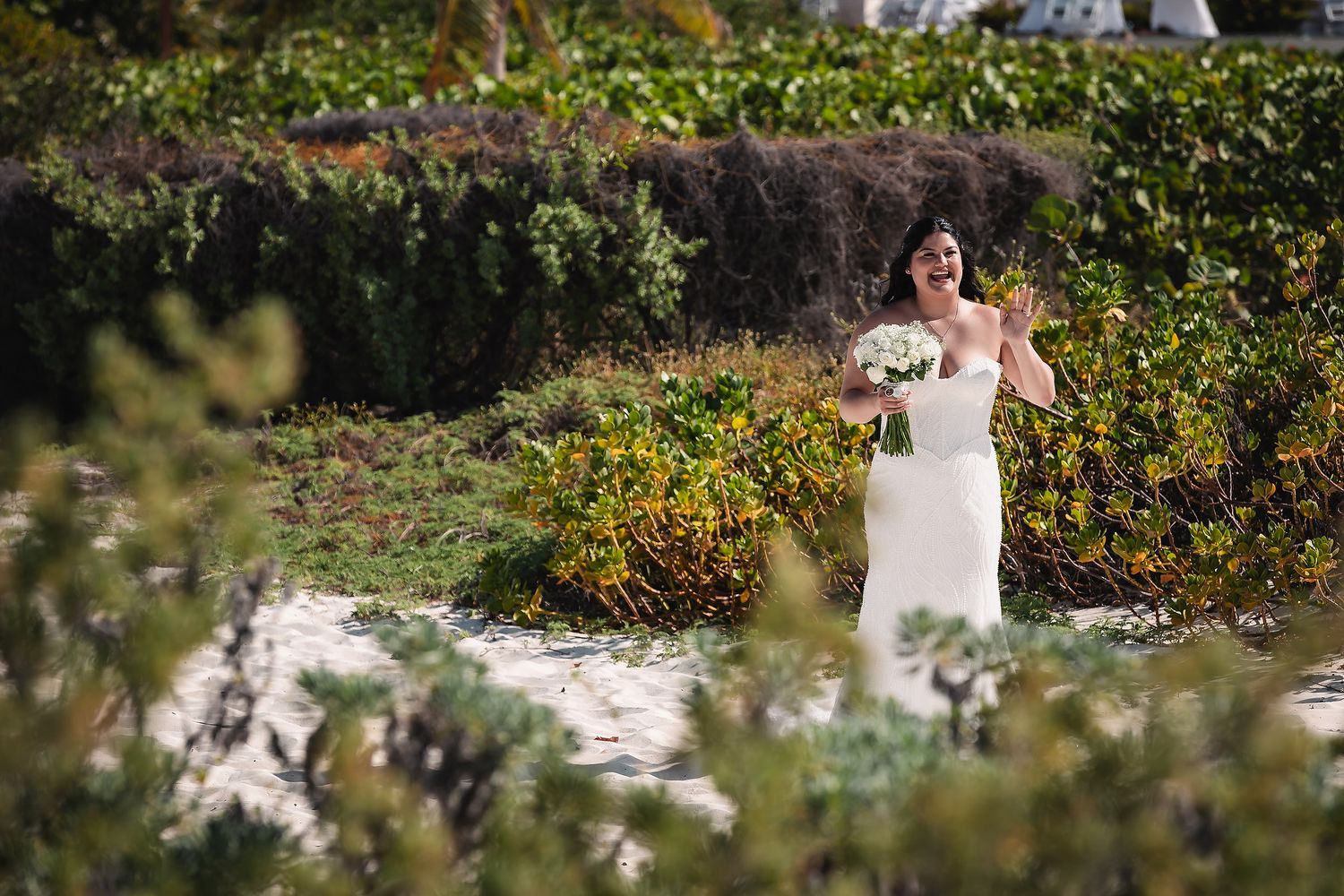 Bride in white gown holding bouquet walking through lush greenery at beach wedding.