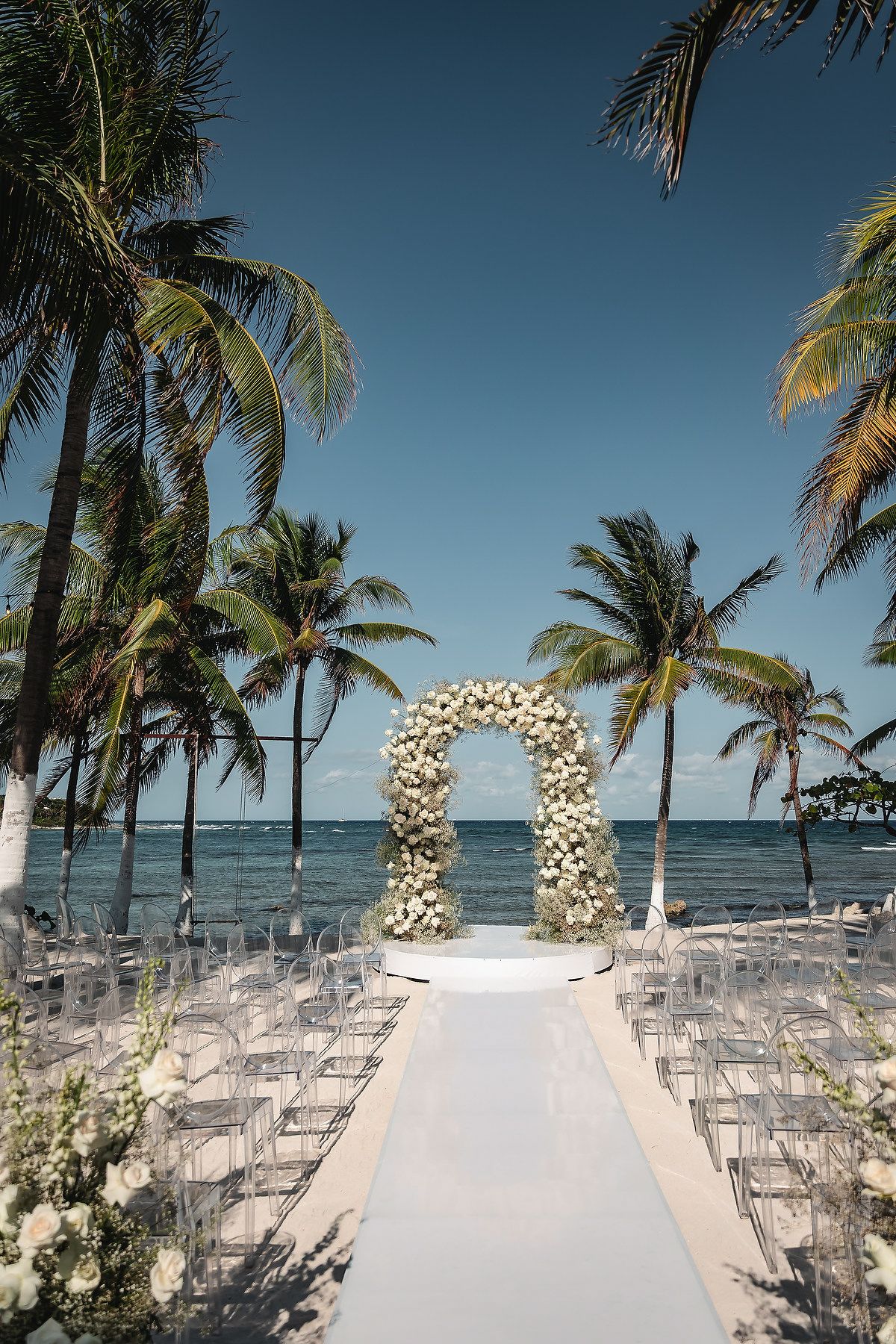 Beachfront wedding ceremony setup with floral arch, white aisle, acrylic chairs, and palm trees overlooking turquoise ocean.