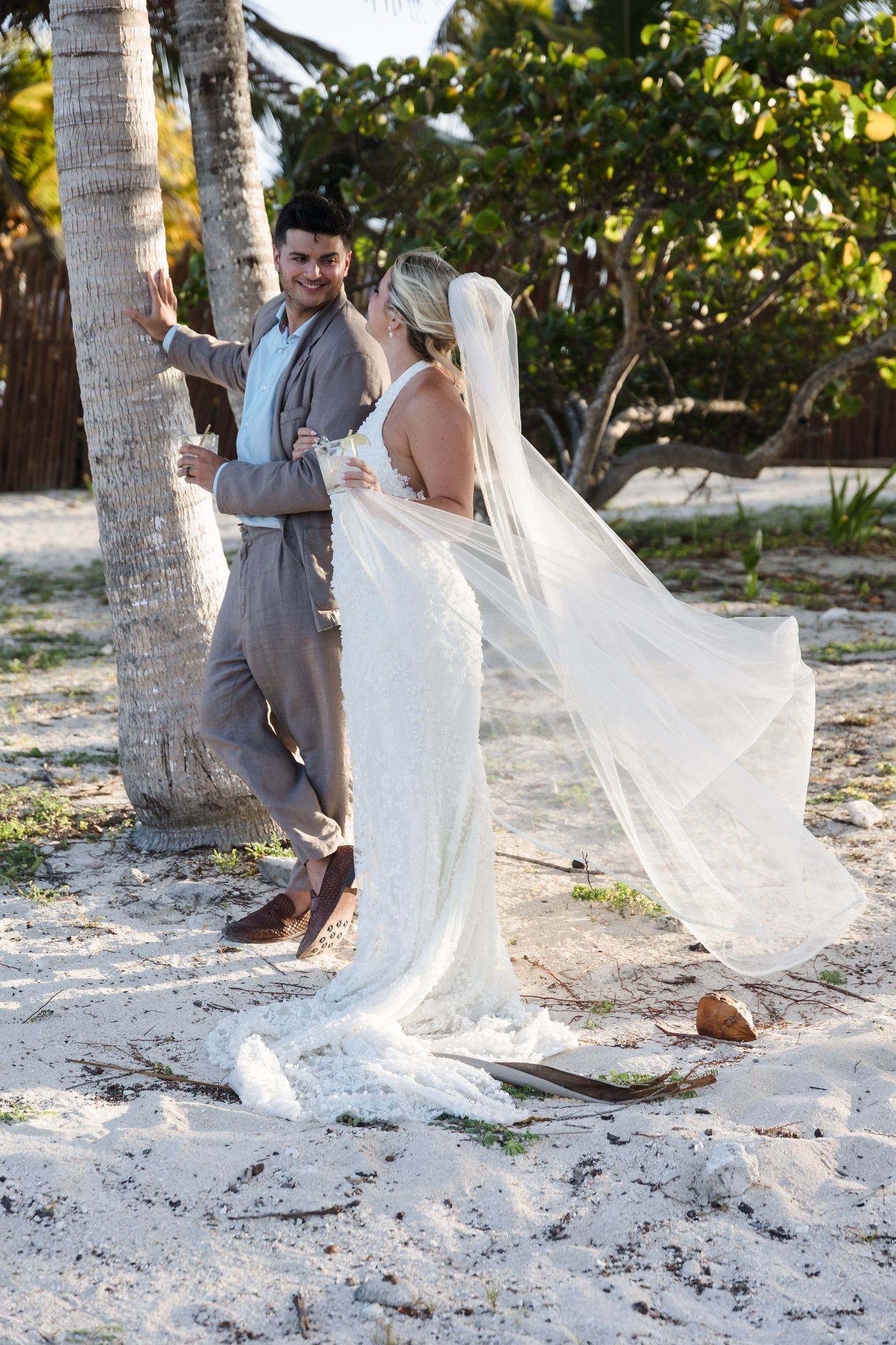 Bride in flowing white gown and veil with groom in tan suit sharing a romantic moment on a tropical beach.