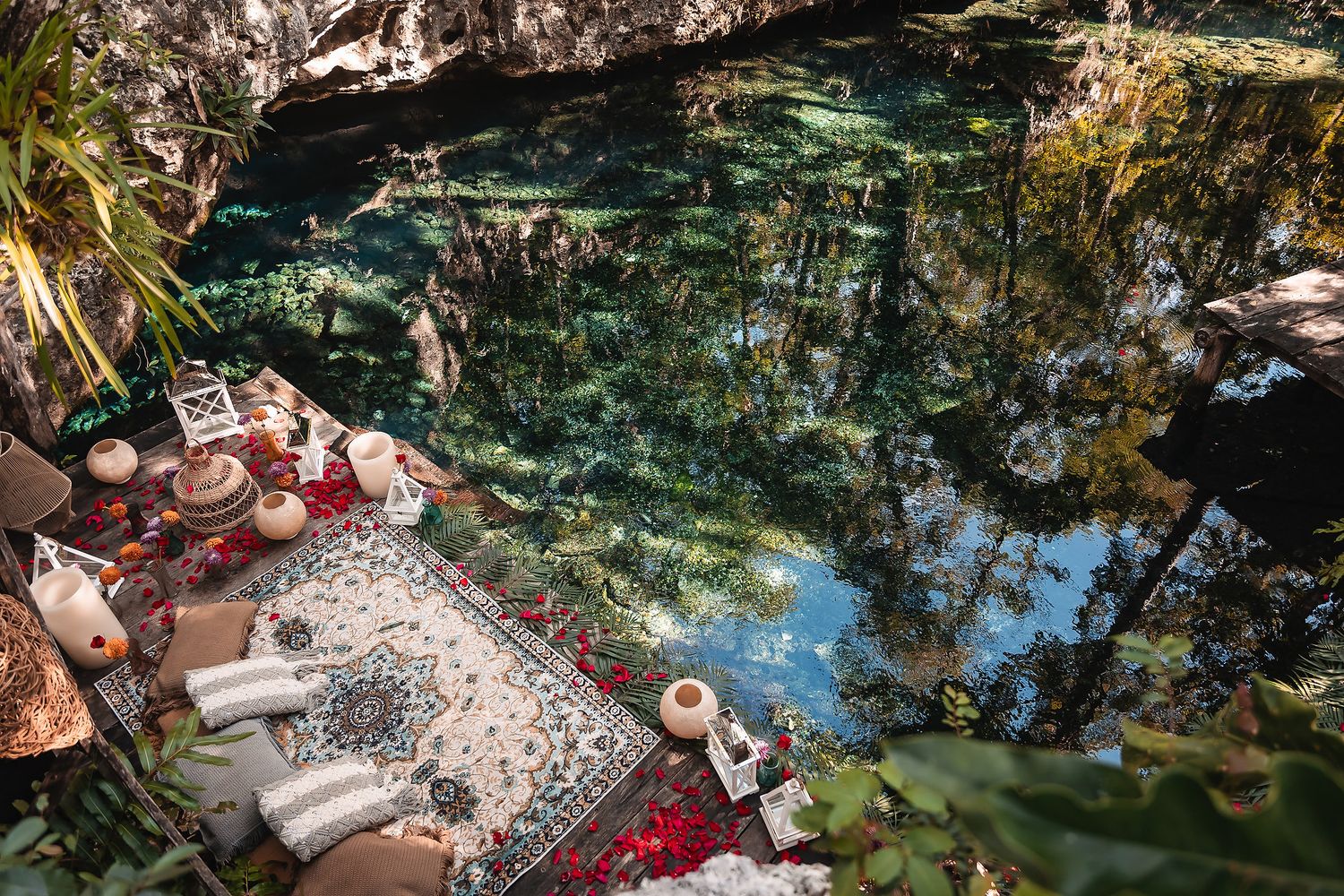 Aerial view of a luxurious cenote setup with ornate rugs, candles, and cushions beside crystal-clear turquoise water.