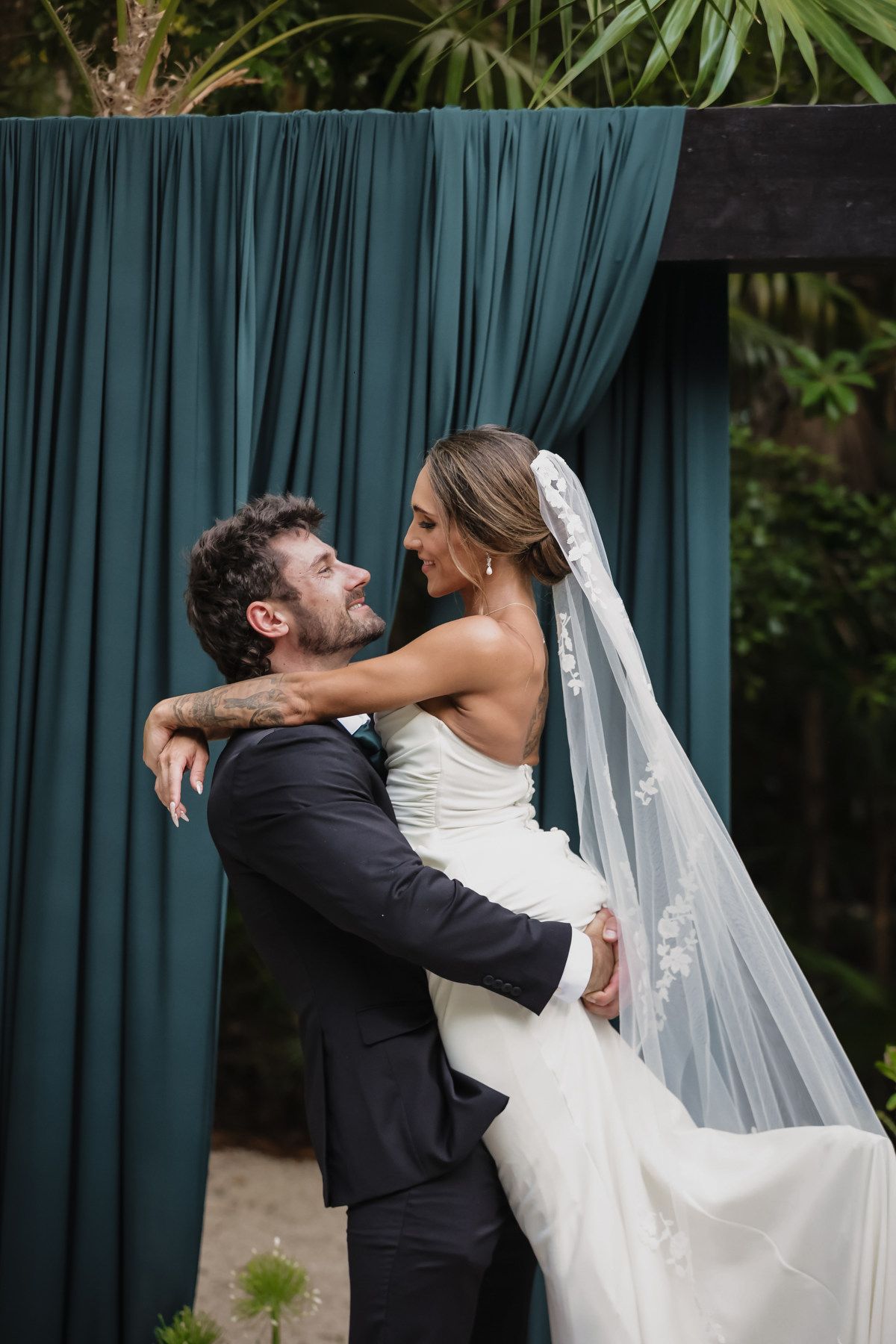 Groom lifts bride in white gown and veil before teal draped wedding backdrop surrounded by lush greenery.