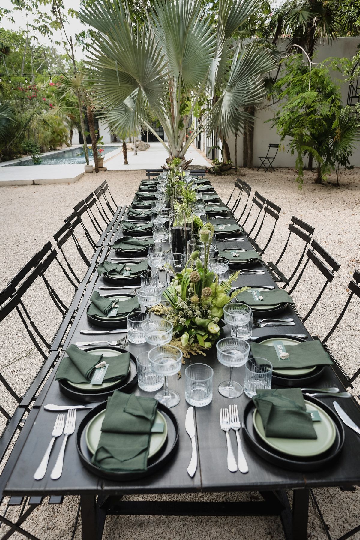 Elegant outdoor dining table set with dark plates, green napkins, crystal glasses, and tropical floral centerpieces on black furniture.