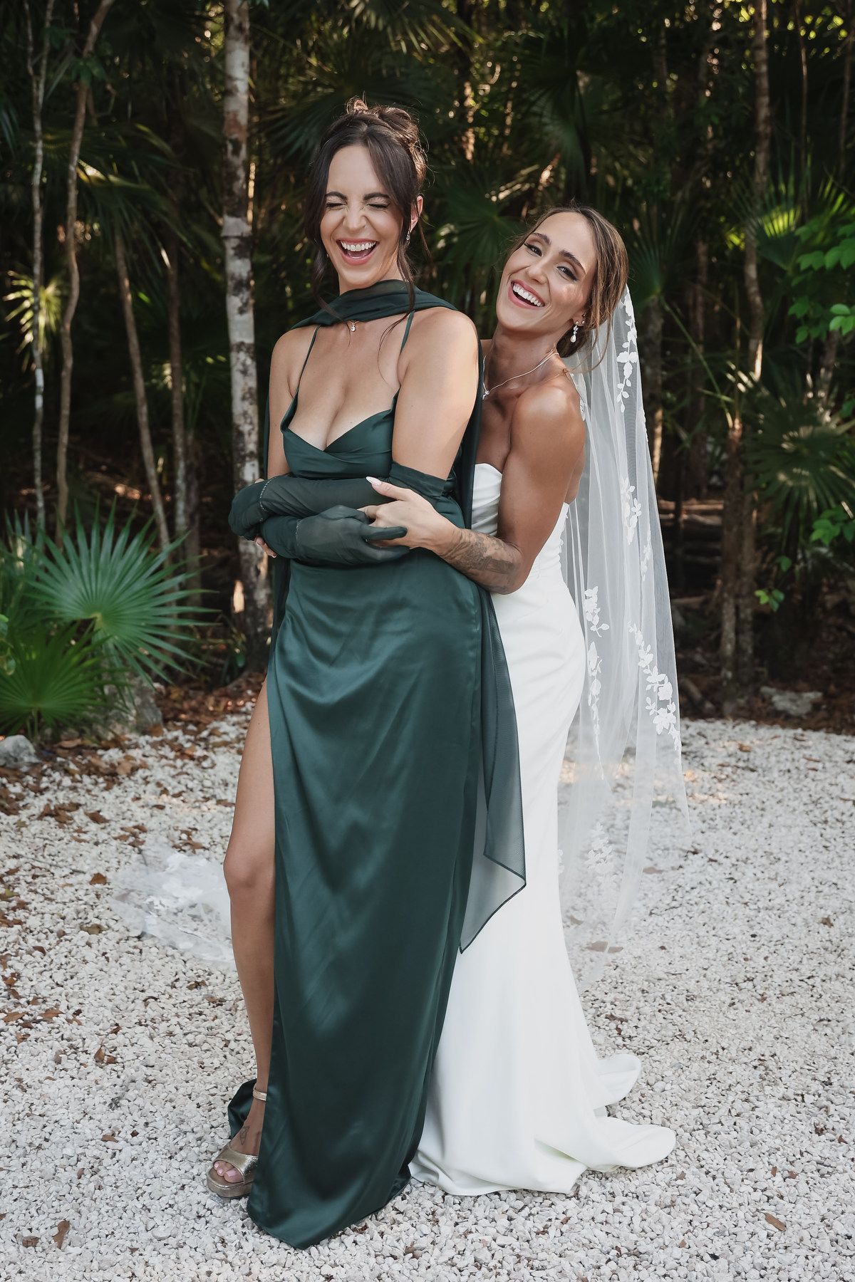 Bride in white gown with veil hugs bridesmaid in elegant green satin dress outdoors among tropical trees.