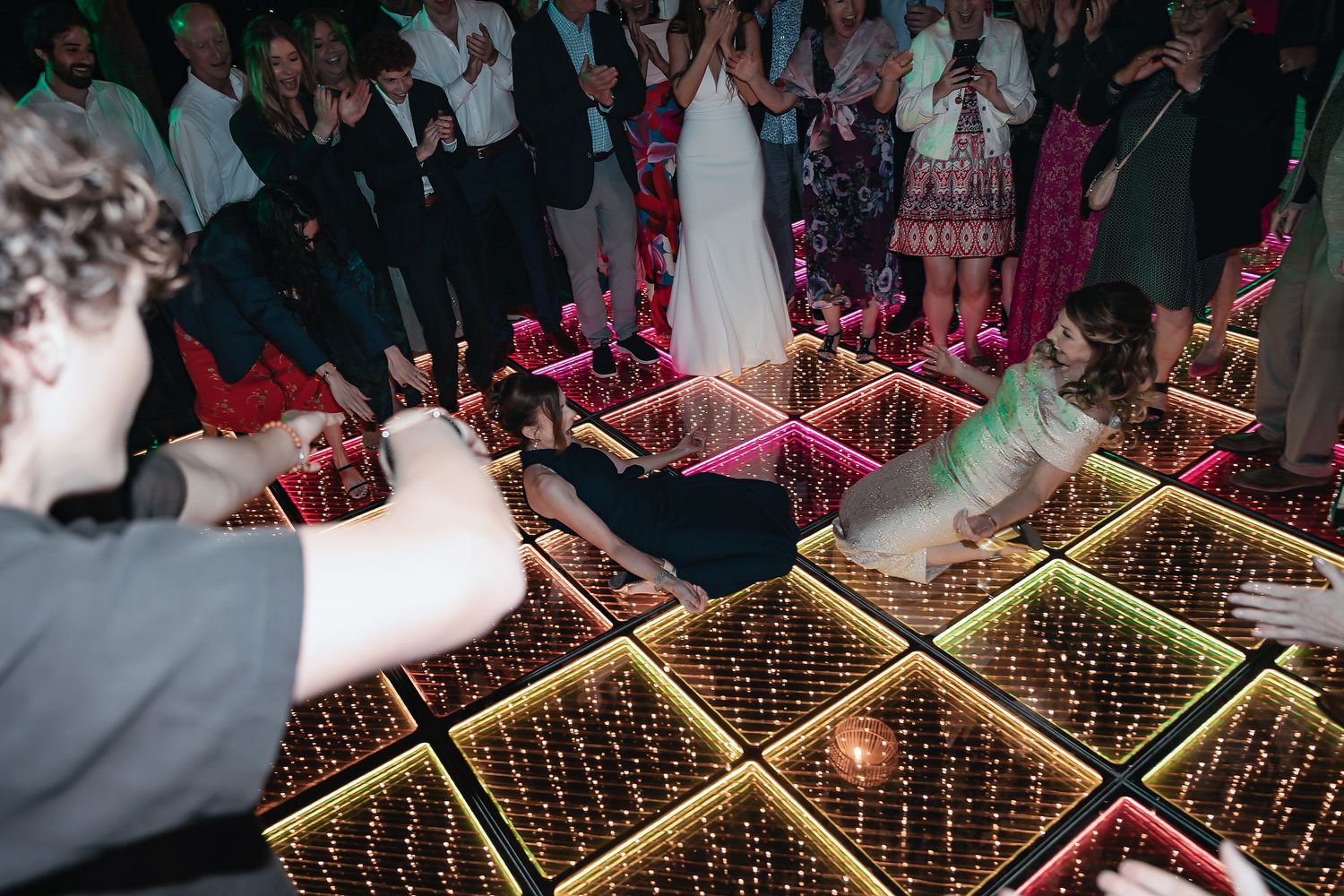 Guests cheer as two people lie on a glowing LED dance floor at a lively wedding reception party.