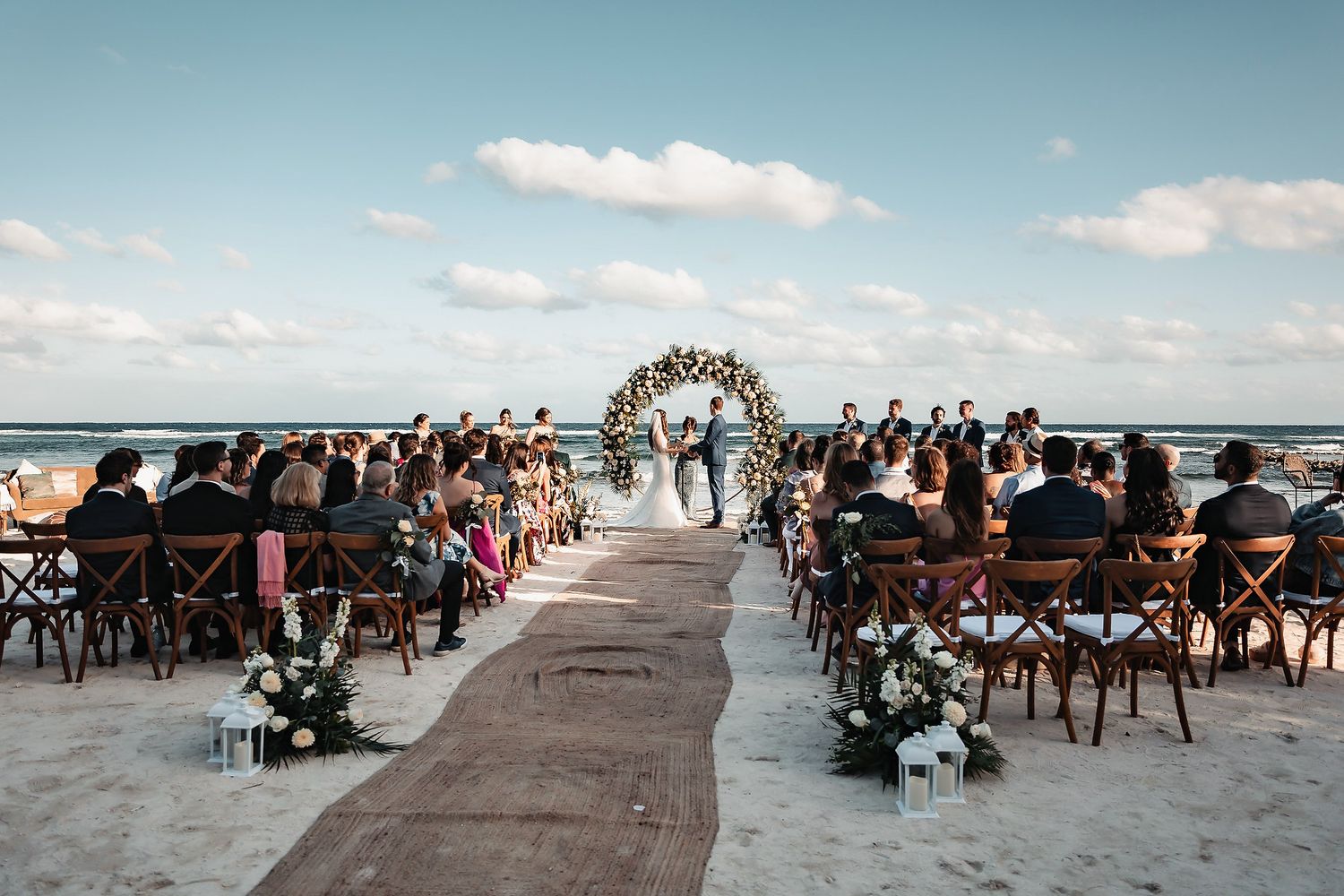 Elegant beach wedding ceremony with guests seated on wooden chairs along a sandy aisle leading to a floral arch by the ocean.