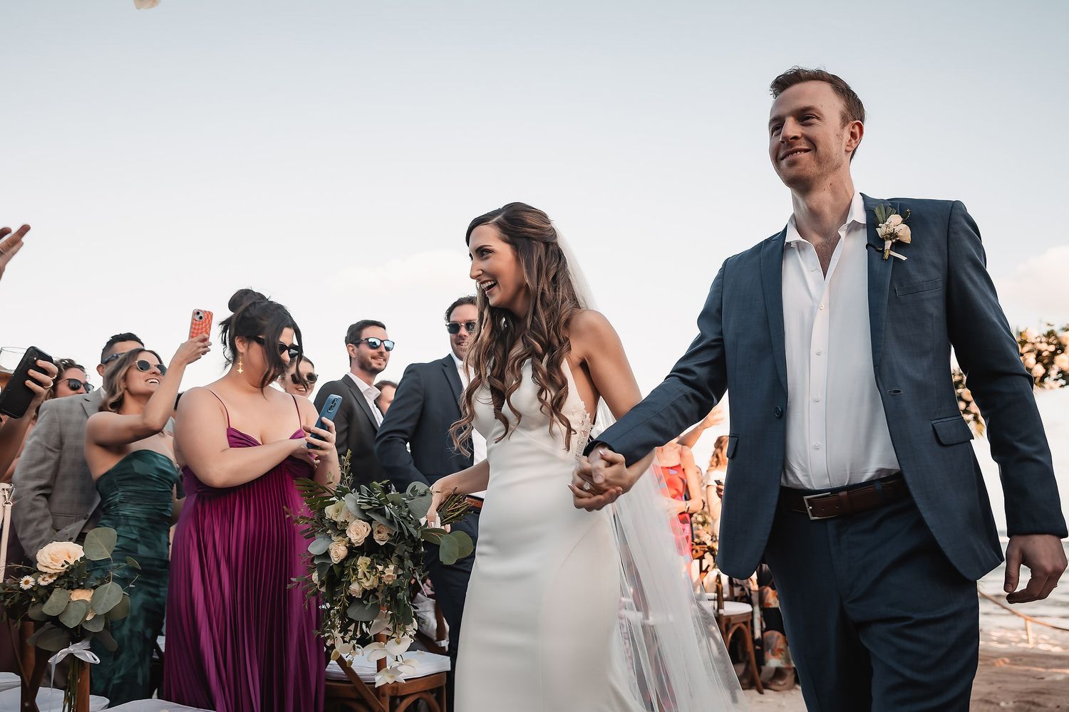 Happy newlyweds walk hand-in-hand at outdoor beach wedding ceremony while guests celebrate around them.
