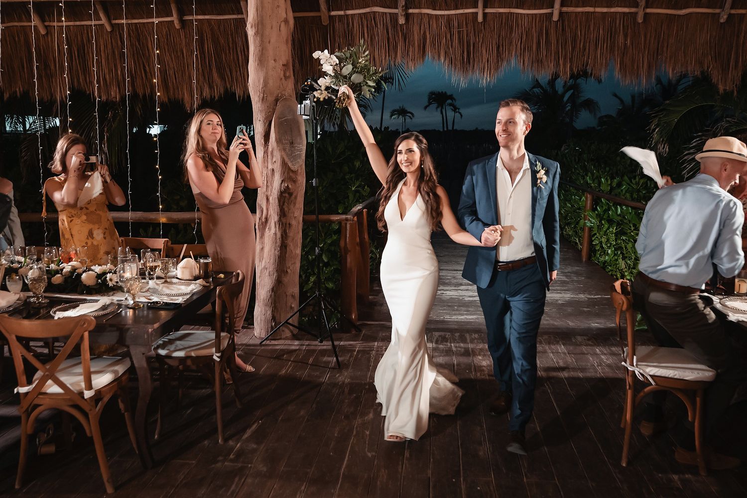 Bride and groom make joyful reception entrance at tropical outdoor wedding venue with thatched roof.