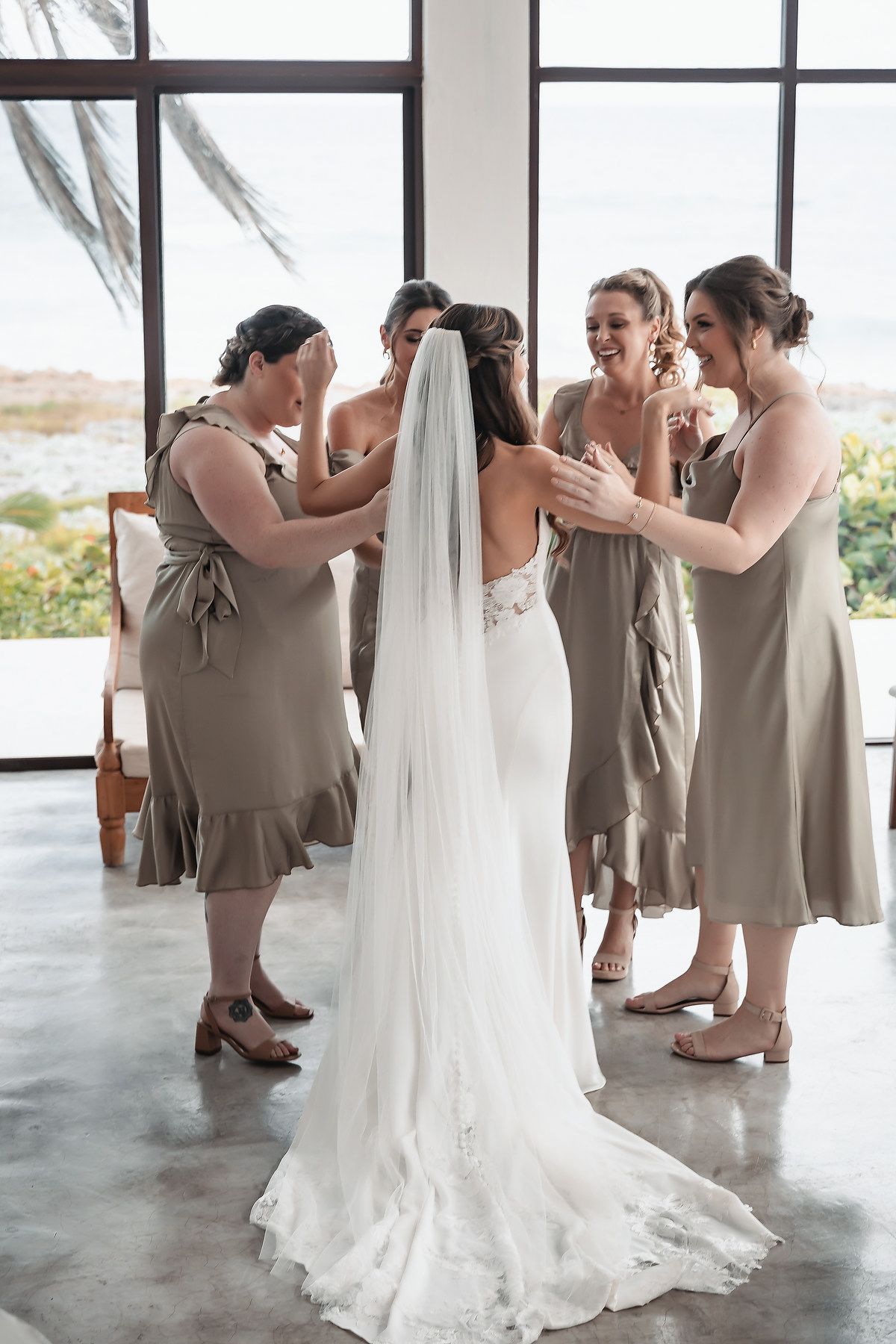 Bride in white gown and veil surrounded by bridesmaids in sage green dresses helping her prepare for wedding.