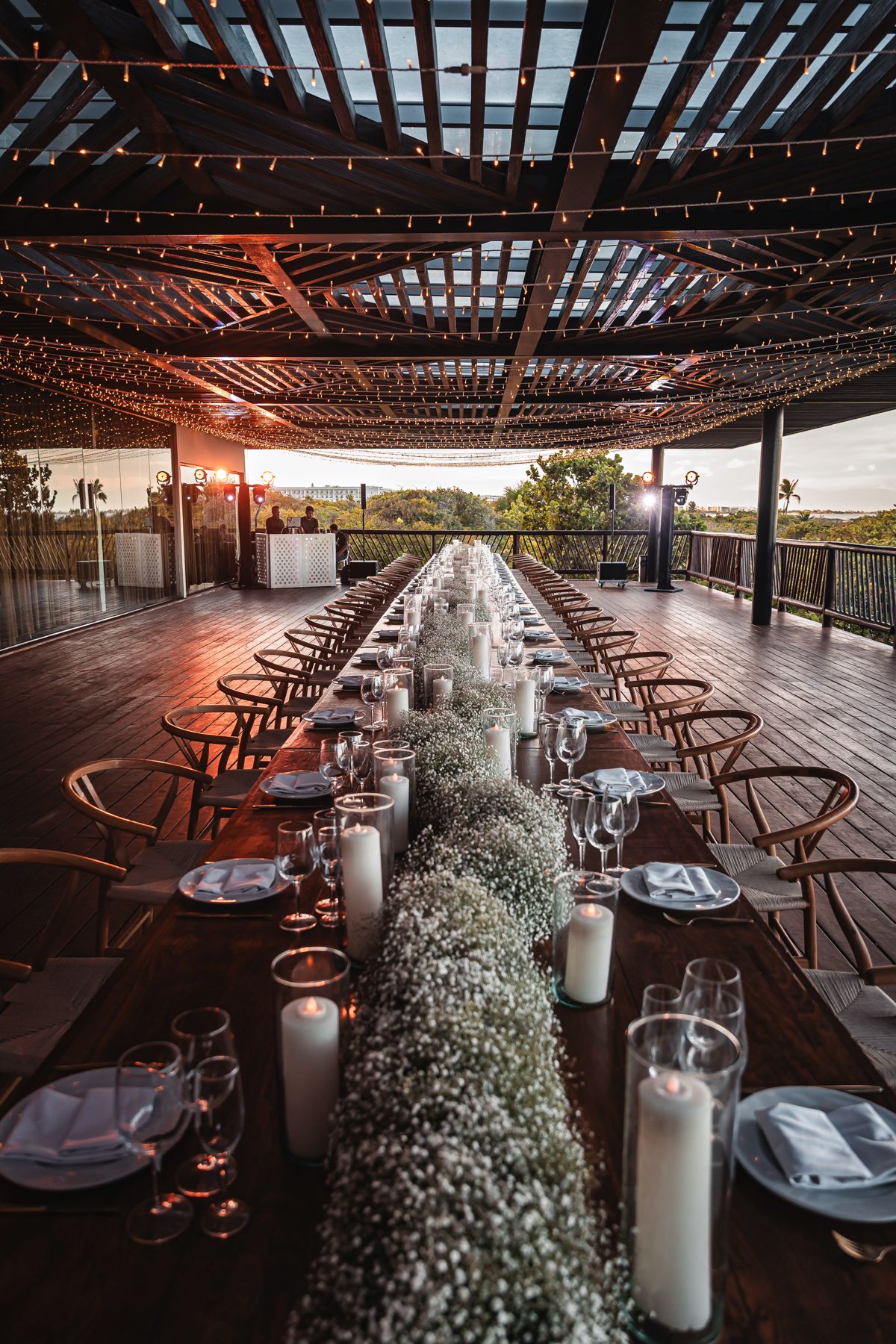 Elegant outdoor wedding reception with long wooden table, white floral runner, candles, and string lights under pergola.